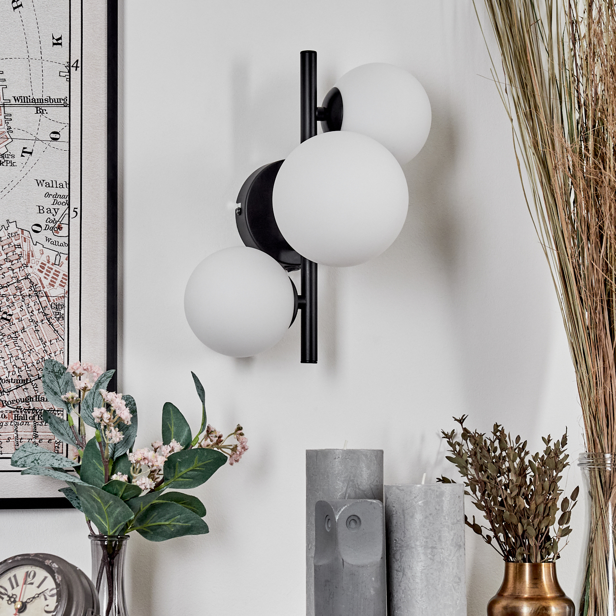 Eine moderne Wandlampe mit drei weißen Kugelleuchten. Daneben sind eine Karte, Blumen, Kerzen und getrocknete Pflanzen zu sehen.
