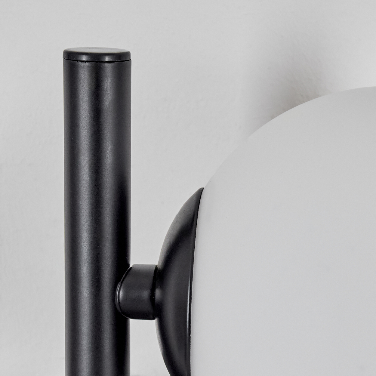 Nahaufnahme einer Lampe. Ein schwarzer Metallständer trägt einen weißen, kugelförmigen Lampenschirm. Der Hintergrund ist eine weiße Wand.