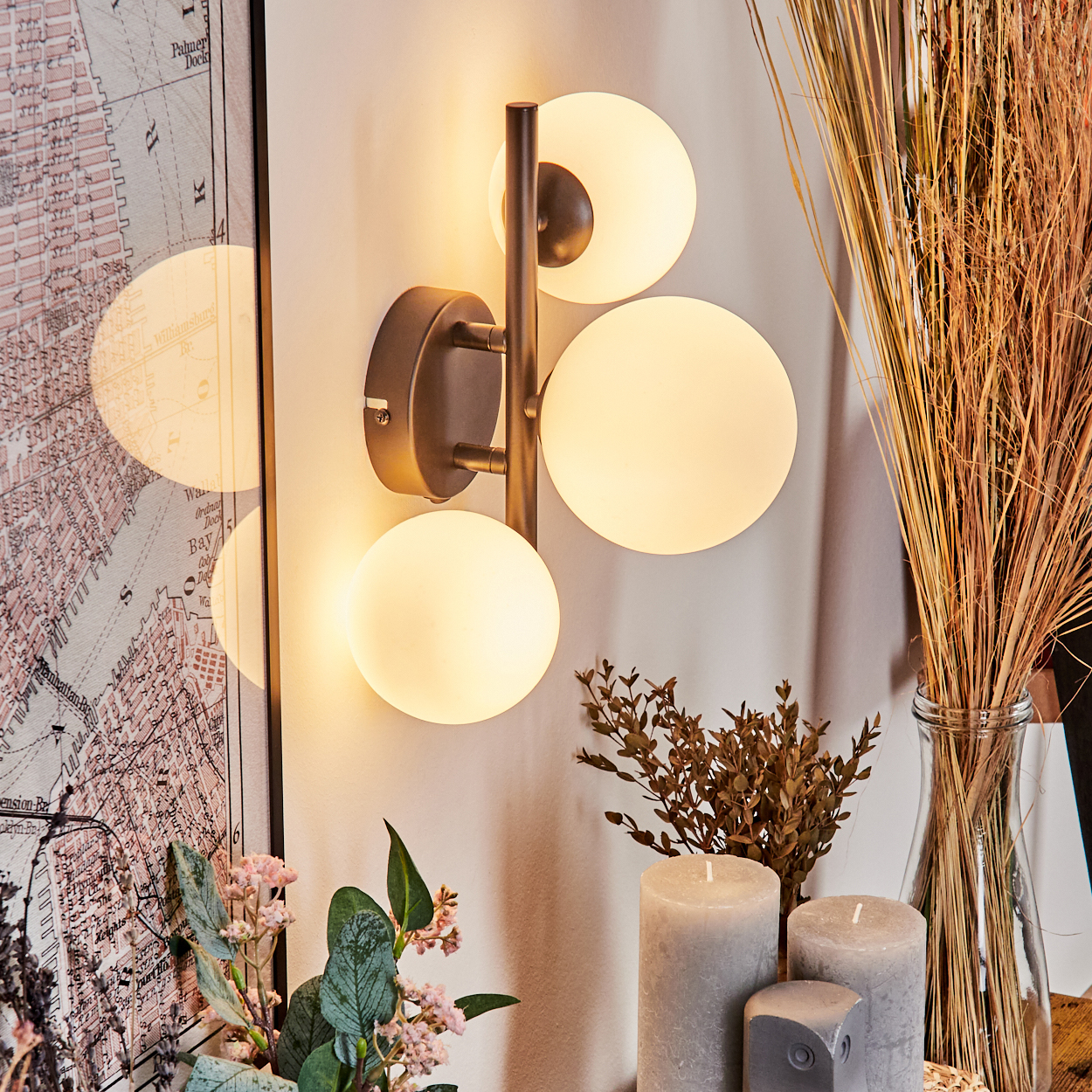 Eine Wandlampe mit drei leuchtenden Kugeln ist an einer weißen Wand montiert. In der Nähe befinden sich Kerzen, trockene Pflanzen und ein Kartendruck.