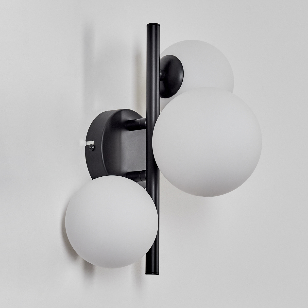 Eine moderne Wandlampe mit drei weißen Kugelleuchten und einem schwarzen Metallrahmen, an einer weißen Wand befestigt.