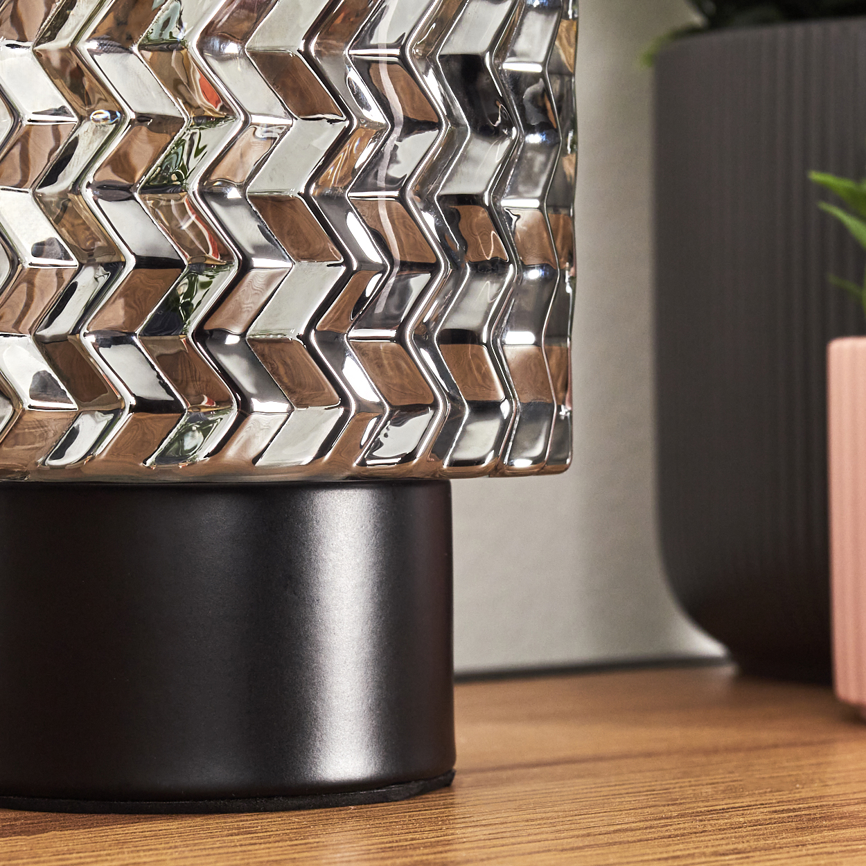 Nahaufnahme einer dekorativen silbernen Vase mit geometrischem Muster, schwarzem Sockel und Holzoberfläche mit unscharfem Hintergrund.