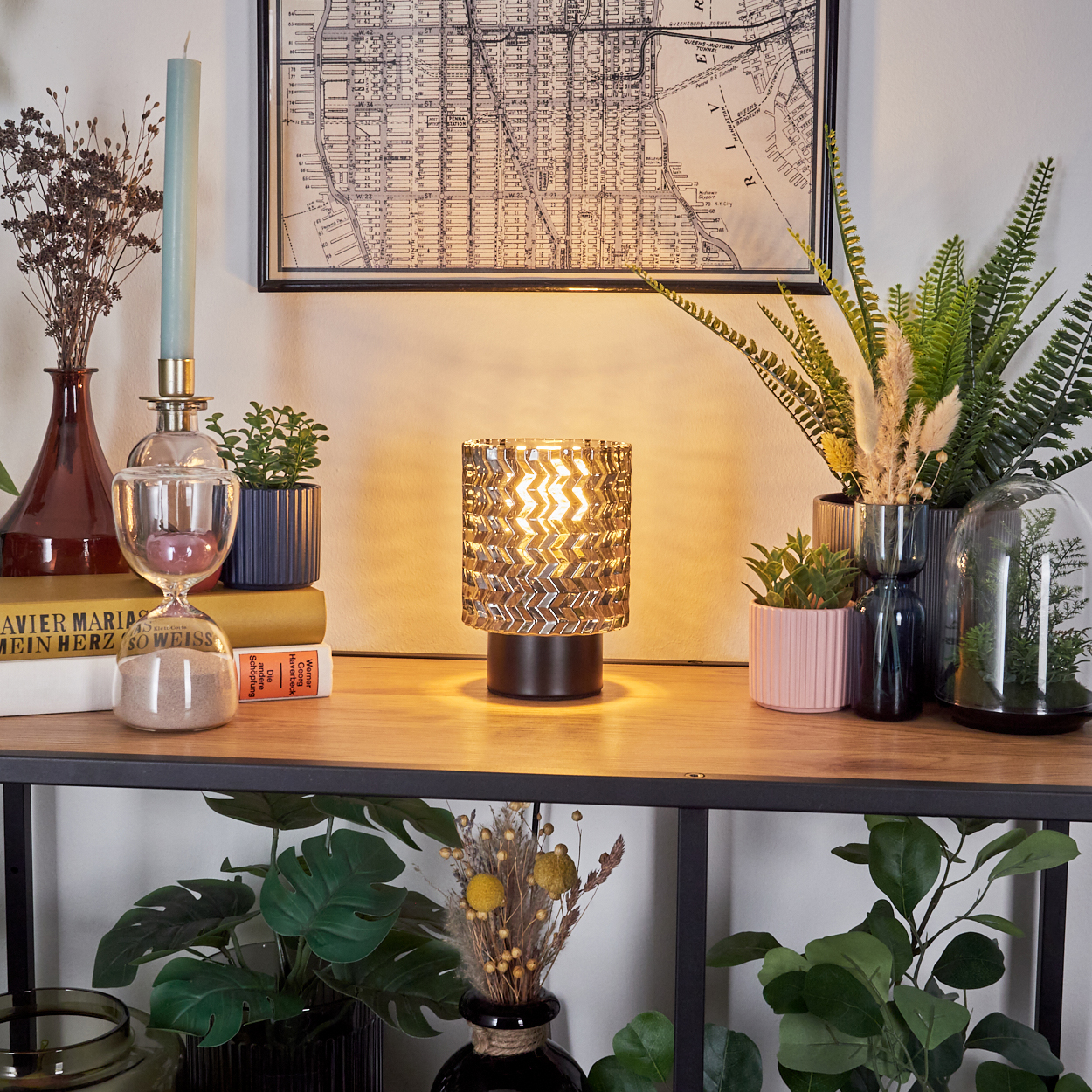Auf einem Tisch stehen Lampe, Pflanzen, Bücher und eine Karte. Die Lampe leuchtet, Pflanzen stehen in Vasen, eine Karte hängt an der Wand.