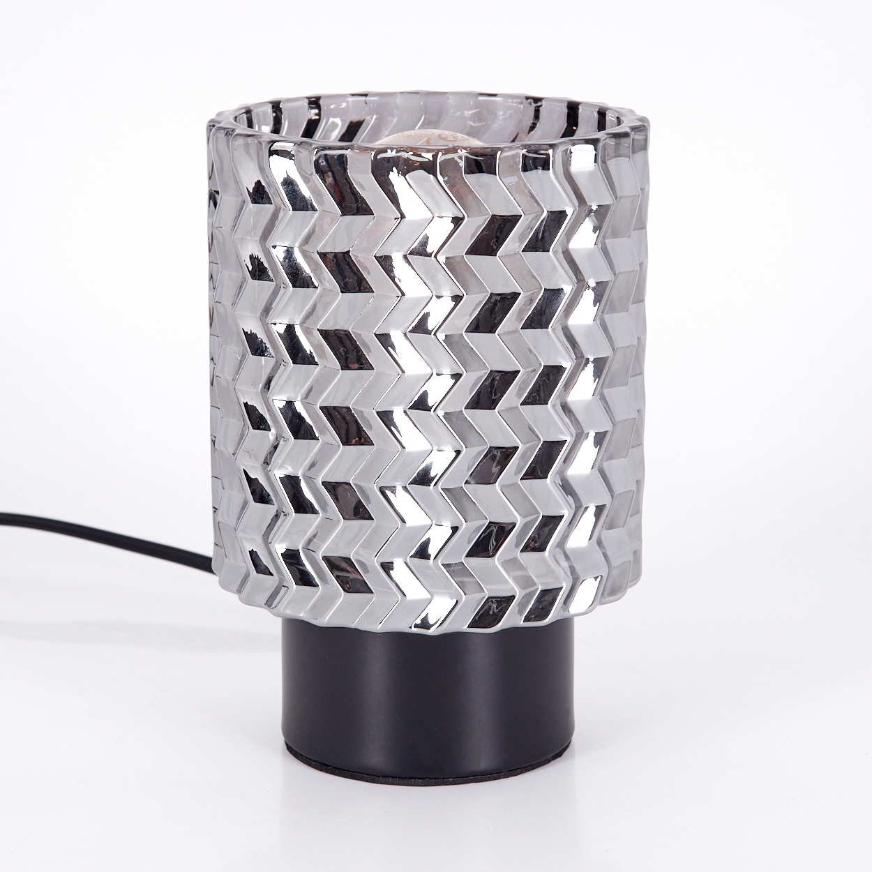 Eine silberne Lampe mit geometrischem Muster auf schwarzem Sockel und schwarzem Kabel. Der Hintergrund ist weiß.