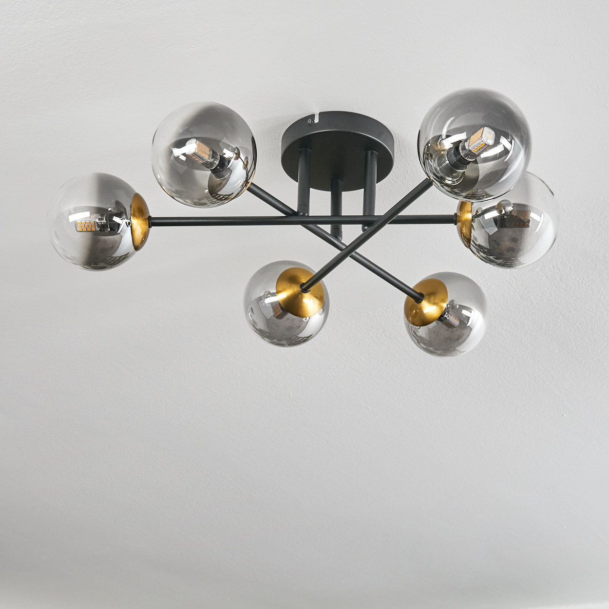 Eine moderne Deckenleuchte mit sechs rauchfarbenen Glaskugeln, schwarzen Armen und goldenen Akzenten. Sie befindet sich an einer weißen Decke.