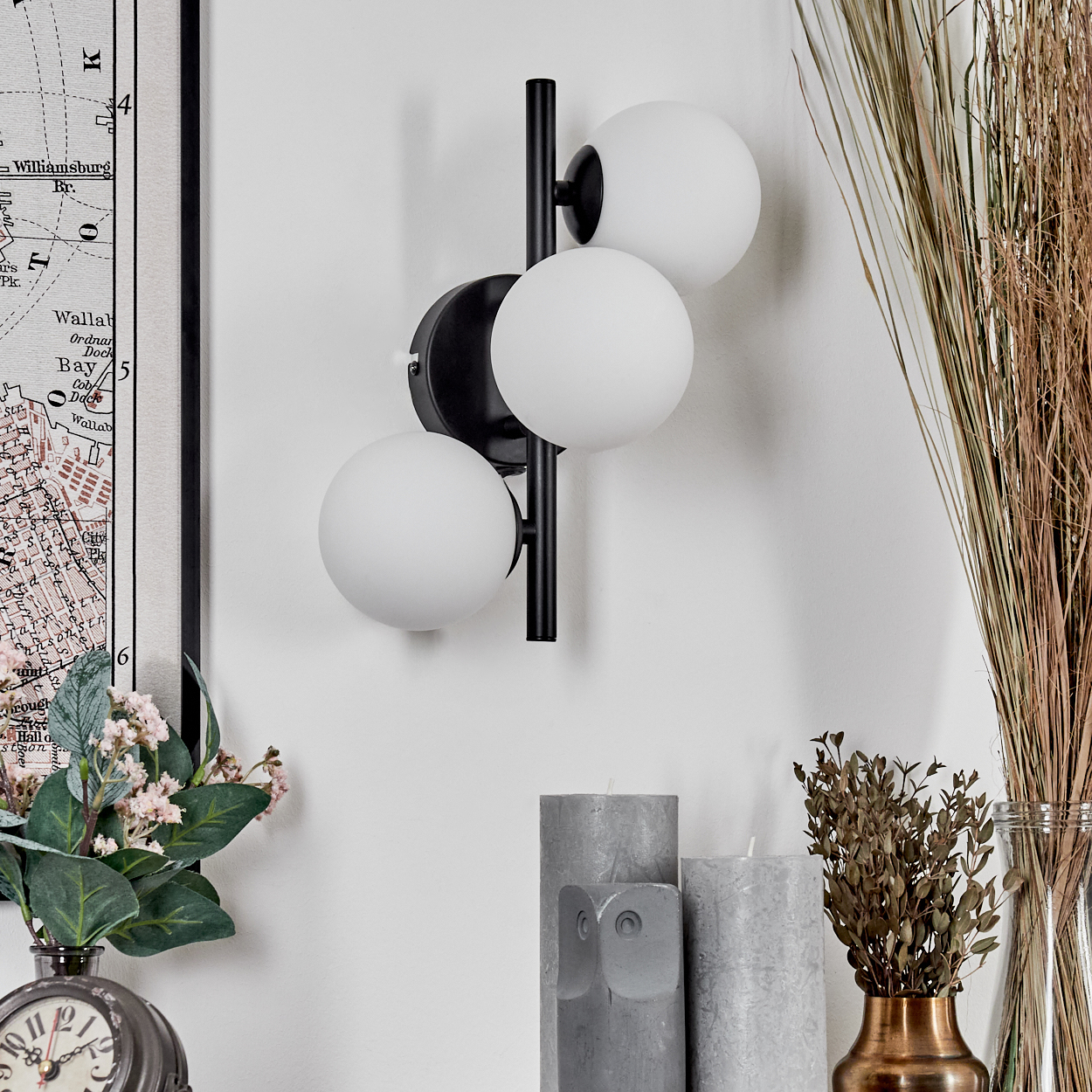 Moderne Wandlampe mit drei weißen Kugelleuchten. Darunter graue Kerzen und eine Vase mit getrockneten Pflanzen. Links eine Karte.