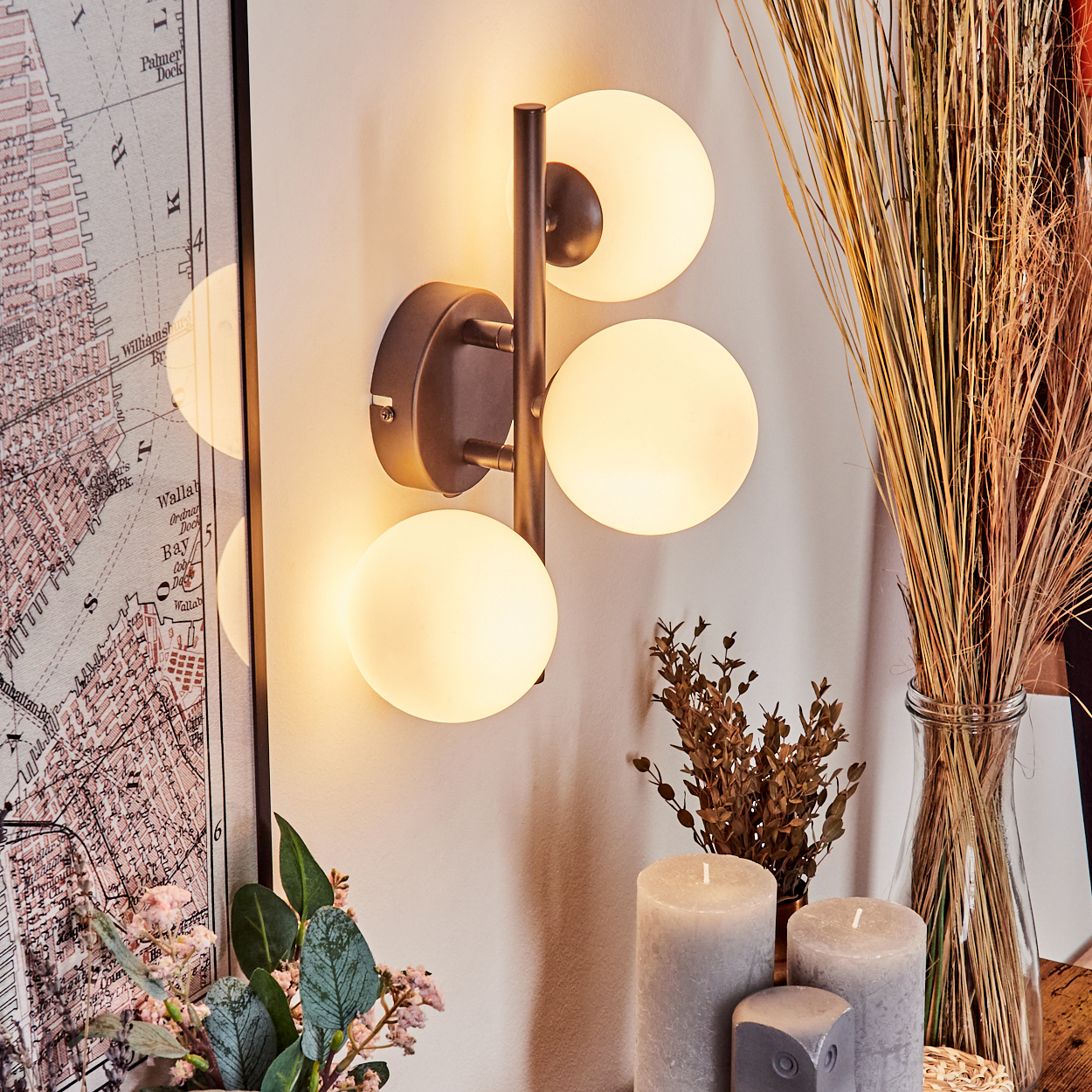 Eine Wandlampe mit drei leuchtenden Kugeln ist an einer hellen Wand montiert. Kerzen, eine Vase mit Gras und eine Karte sind im Bild.