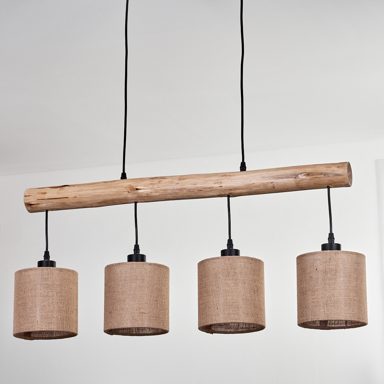 Ein Holzbalken mit vier Hängelampen. Jede Lampe hat einen zylindrischen Juteschirmen und ein schwarzes Kabel. Der Hintergrund ist eine weiße Wand.