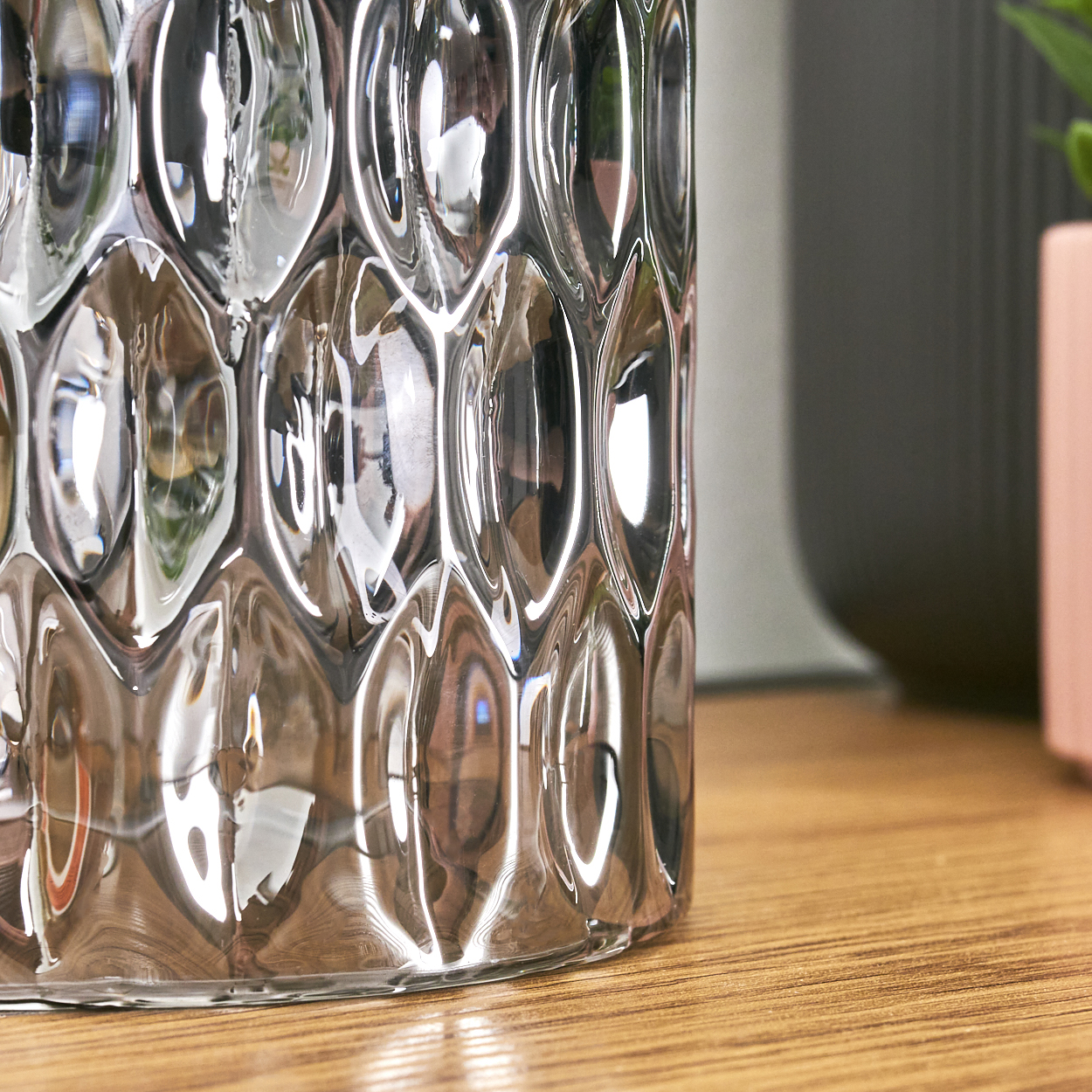Nahaufnahme eines durchsichtigen, strukturierten Glasgefäßes, das einen Raum reflektiert. Eine Holzoberfläche trägt es. Der Hintergrund hat ein graues Objekt und einen rosa Topf.