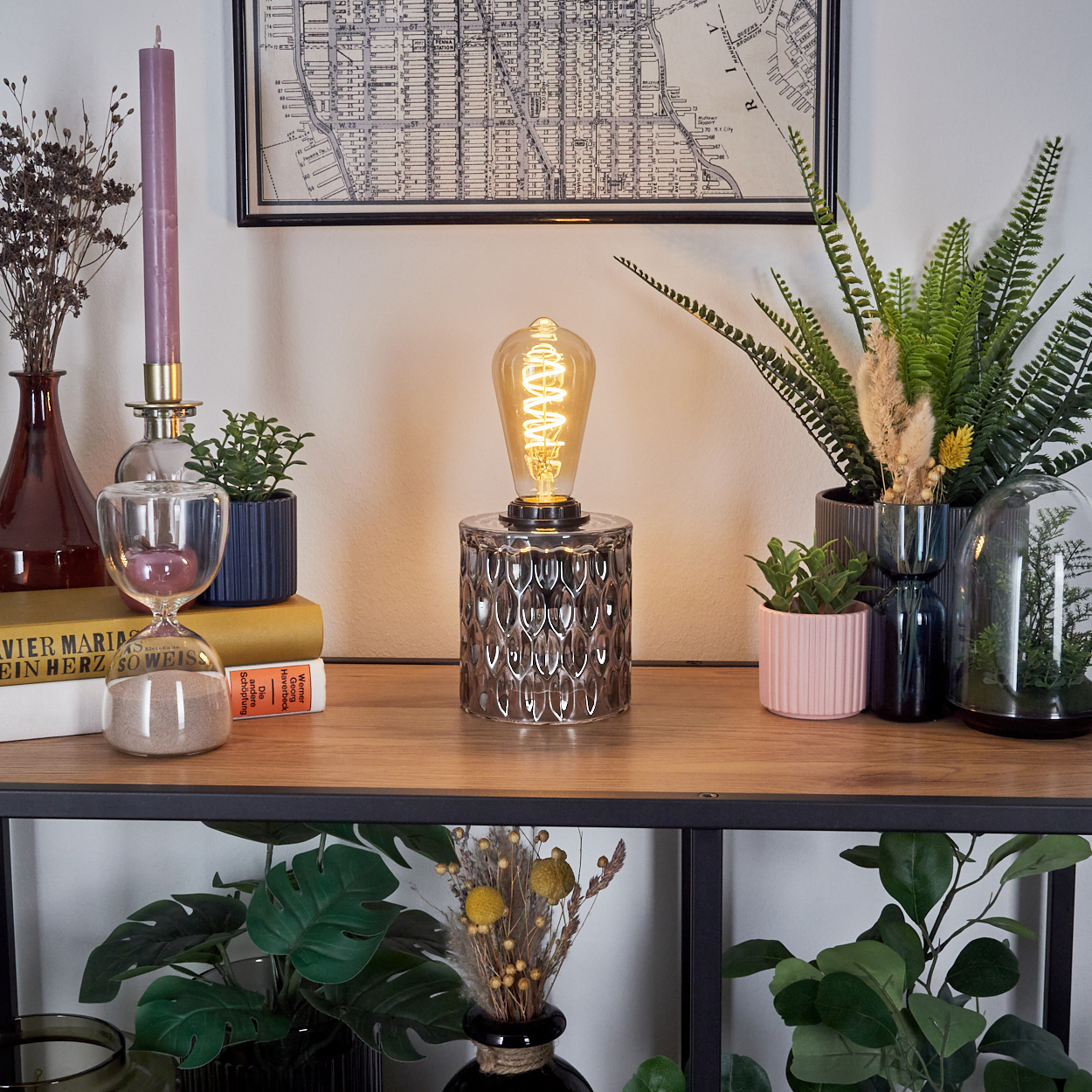 Ein Holzregal zeigt eine Lampe, Pflanzen, eine Sanduhr, eine Kerze und Bücher. Im Hintergrund hängt eine Karte. Die Szene ist drinnen.