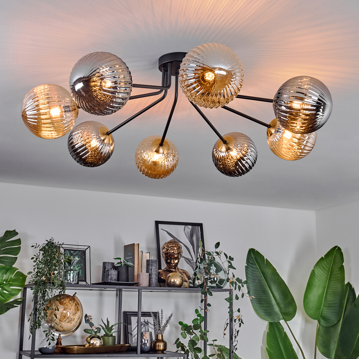 Deckenleuchte mit mehreren Glaskugeln. Darunter ein Regal mit Pflanzen, einem Globus und Kunstwerken. Die Szene ist drinnen und gut beleuchtet.