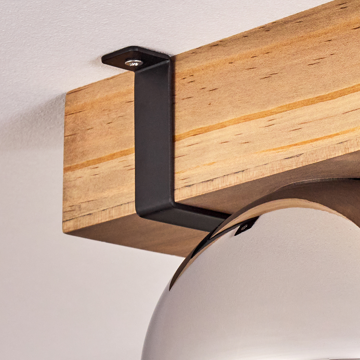 Ein Holzbalken wird von einer schwarzen Metallhalterung gehalten, die an der Decke befestigt ist. Eine weiße, gebogene Lampe befindet sich unter dem Balken.