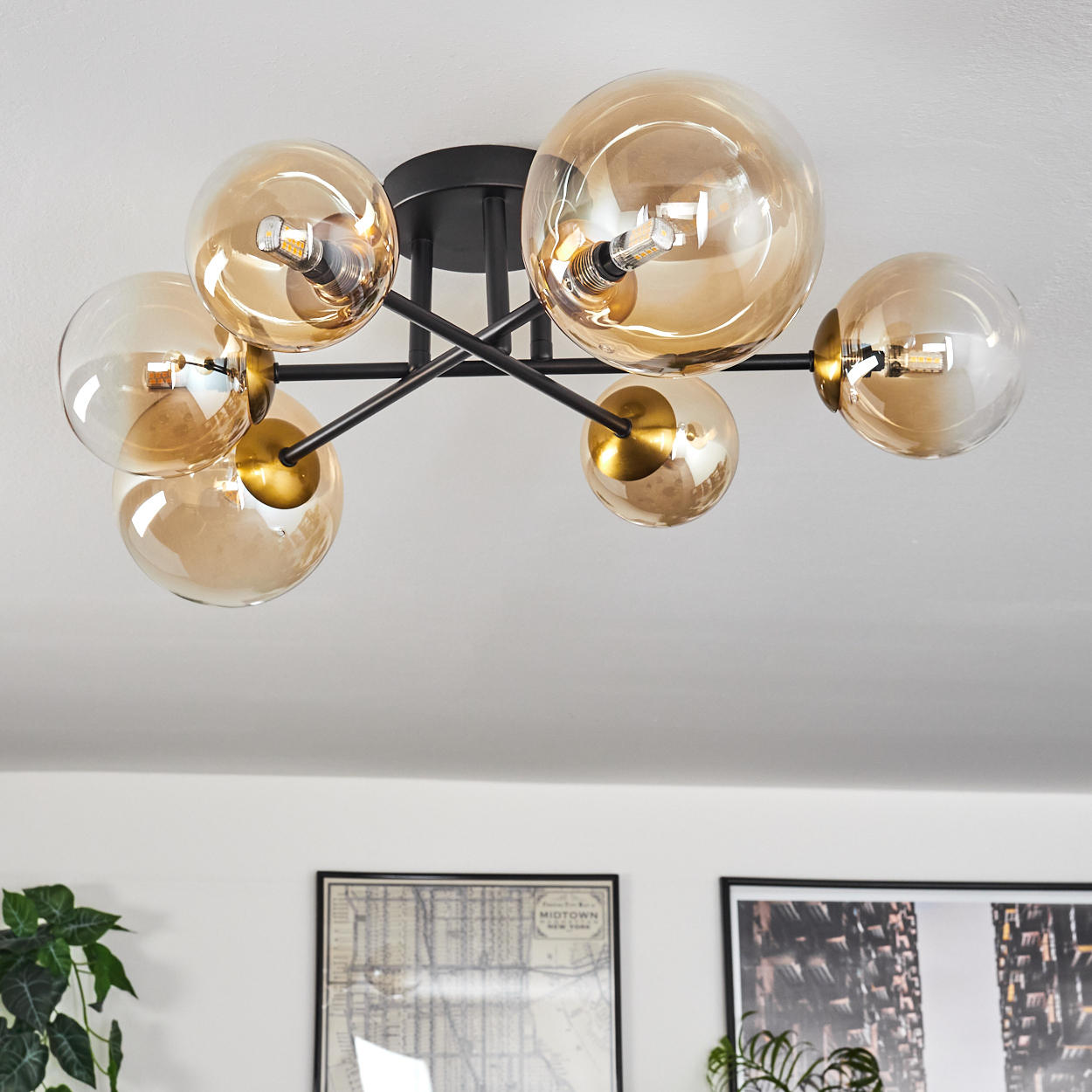 Eine moderne Deckenleuchte mit sechs bernsteinfarbenen Glaskugelschirmen. Schwarze Metallarme und -befestigungen. Dekorative Kunst und Pflanze im Hintergrund.