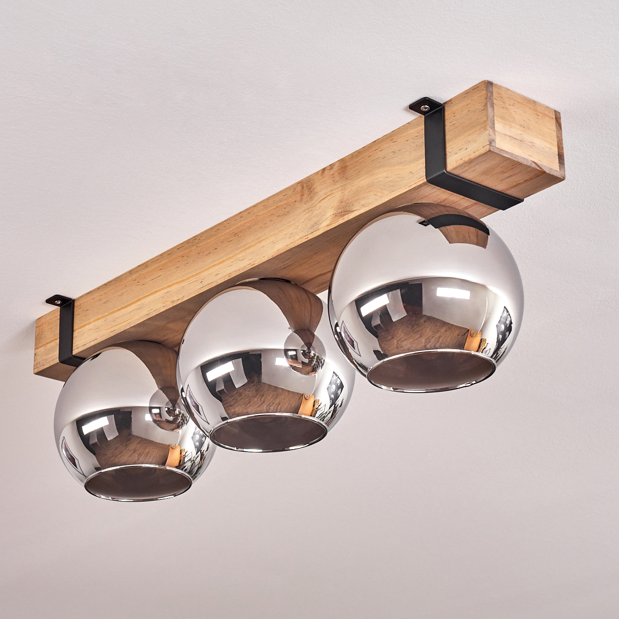 Eine Deckenlampe mit drei verchromten Kugelleuchten, die an einem Holzbalken befestigt sind, der von schwarzen Metallbügeln getragen wird.