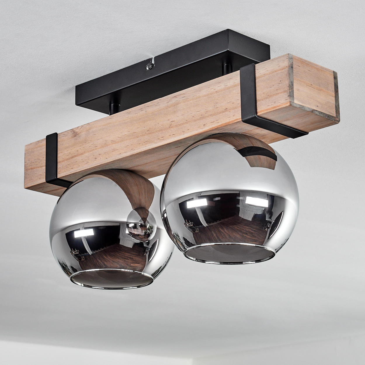 Eine Deckenlampe mit zwei verchromten Kugelleuchten, die an einem Holzbalken hängen. Der Balken wird von schwarzem Metall getragen. Weiße Decke.