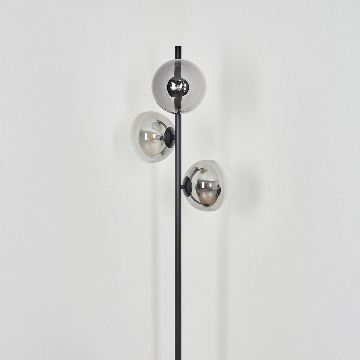 Eine moderne schwarze Lampe mit drei kugelförmigen Glasschirmen. Die Schirme sind klar und reflektieren die Umgebung. Die Lampe steht vor einer weißen Wand.