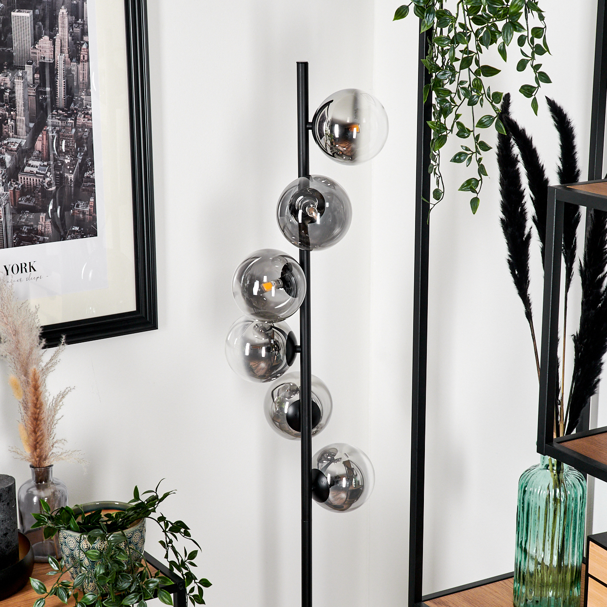Eine moderne Stehlampe mit sechs runden Glasschirmen. Im Hintergrund ein gerahmtes Stadtbild und dekorative Pflanzen.
