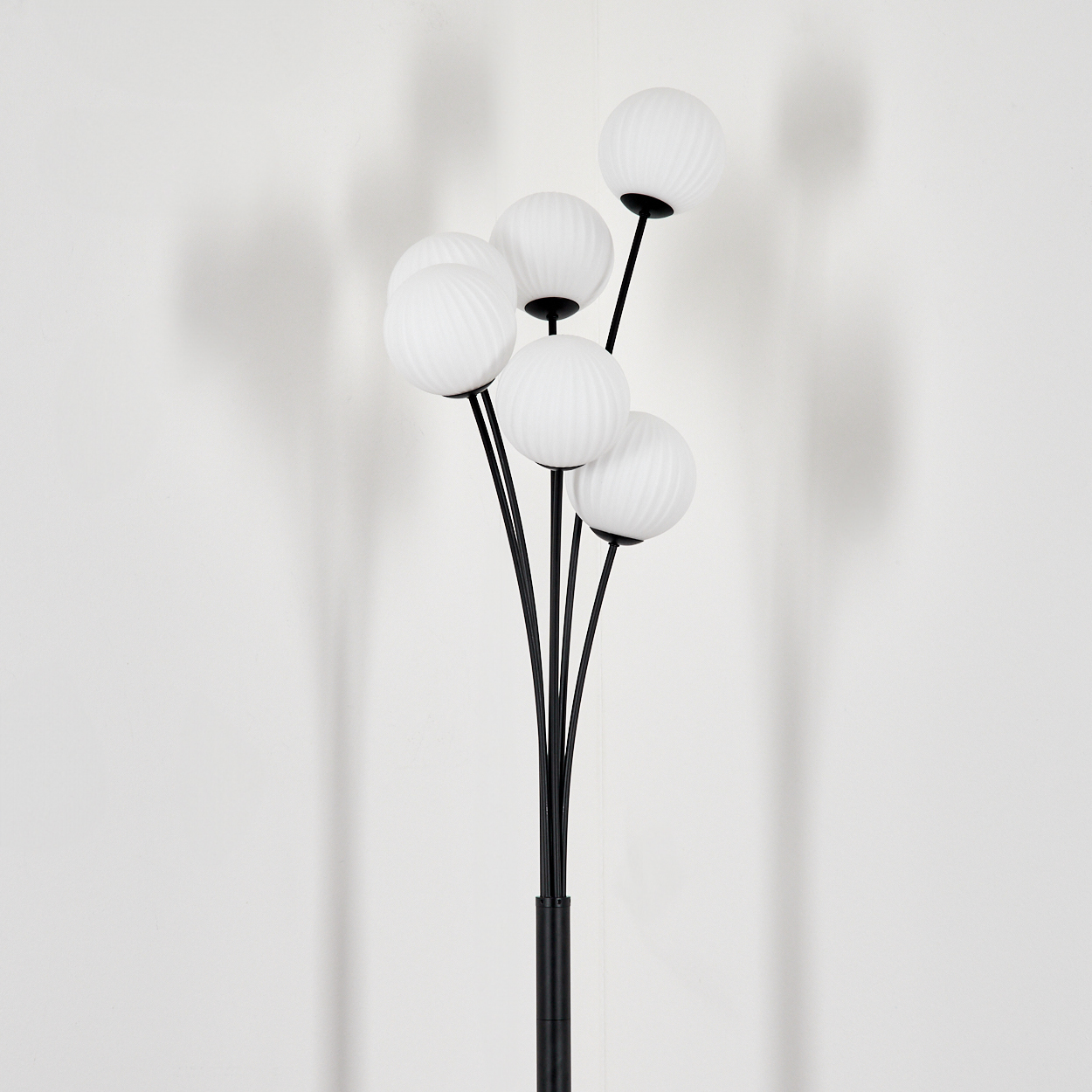 Eine moderne Stehlampe mit fünf weißen Kugelschirmen auf schwarzen Stielen. Der Hintergrund ist eine schlichte weiße Wand mit weichen Schatten.