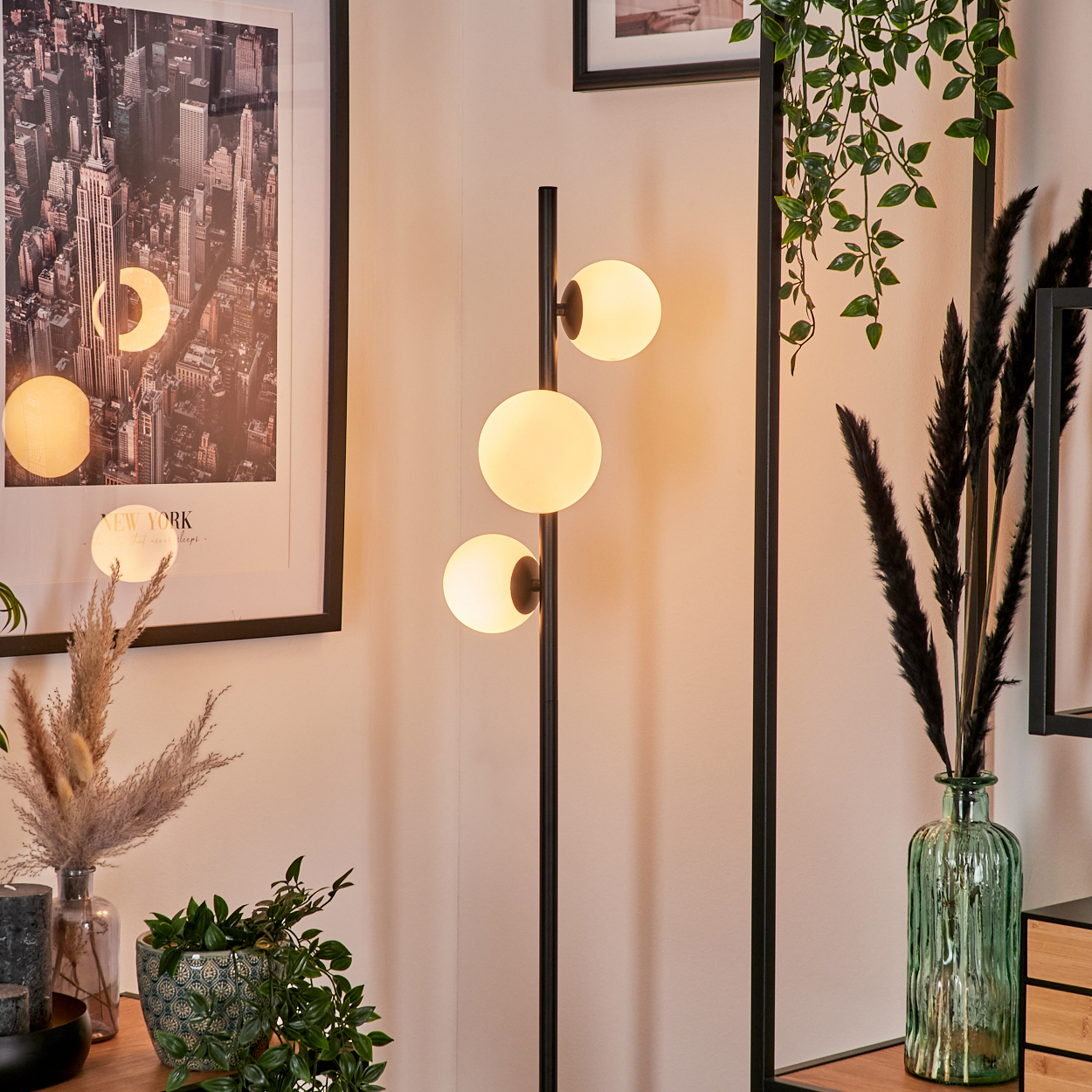 Eine Stehlampe mit drei Kugeln leuchtet. An der Wand hängt ein New York-Poster und im Vordergrund Pflanzen.