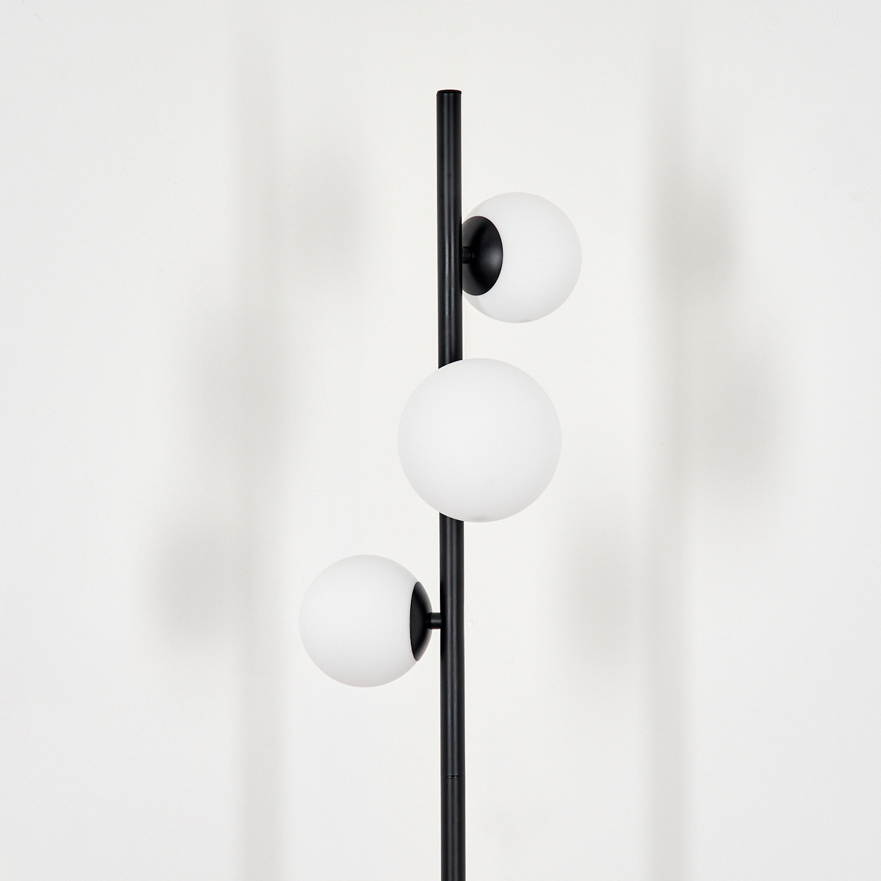 Eine schwarze Stehlampe mit drei kugelförmigen weißen Lichtern. Der Hintergrund ist eine schlichte weiße Wand. Die Lampe steht mittig.
