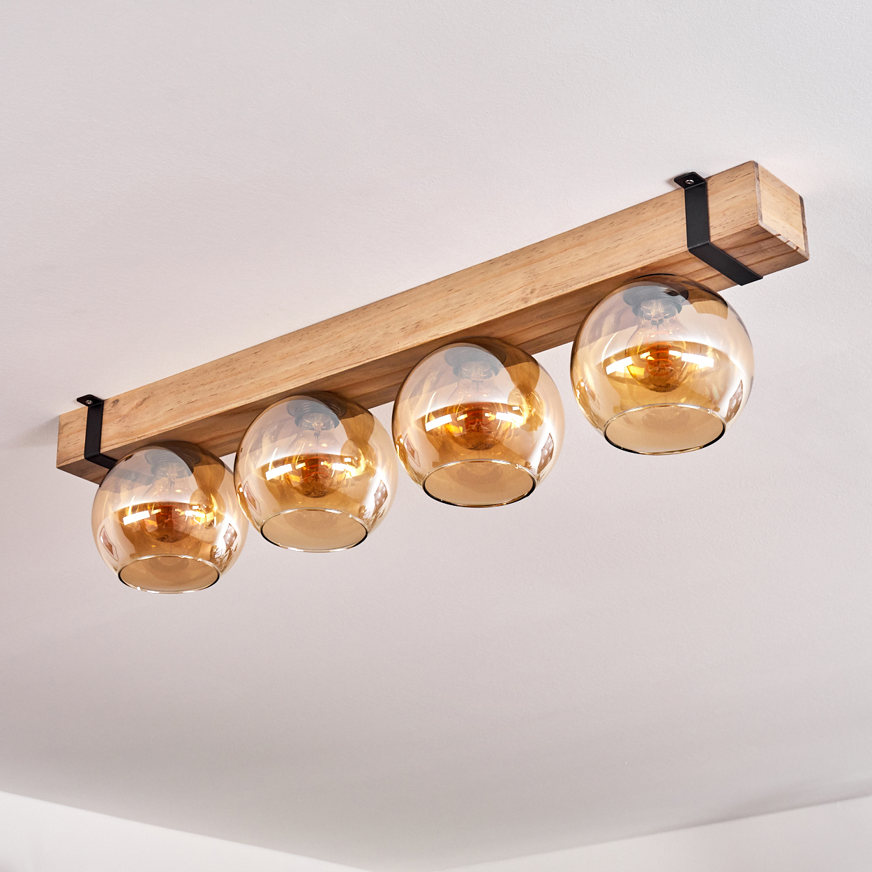 Eine Deckenleuchte aus Holzbalken mit vier bernsteinfarbenen Glaskugelschirmen. Schwarze Metallhalterungen stützen die Leuchte. Der Hintergrund ist eine weiße Decke.