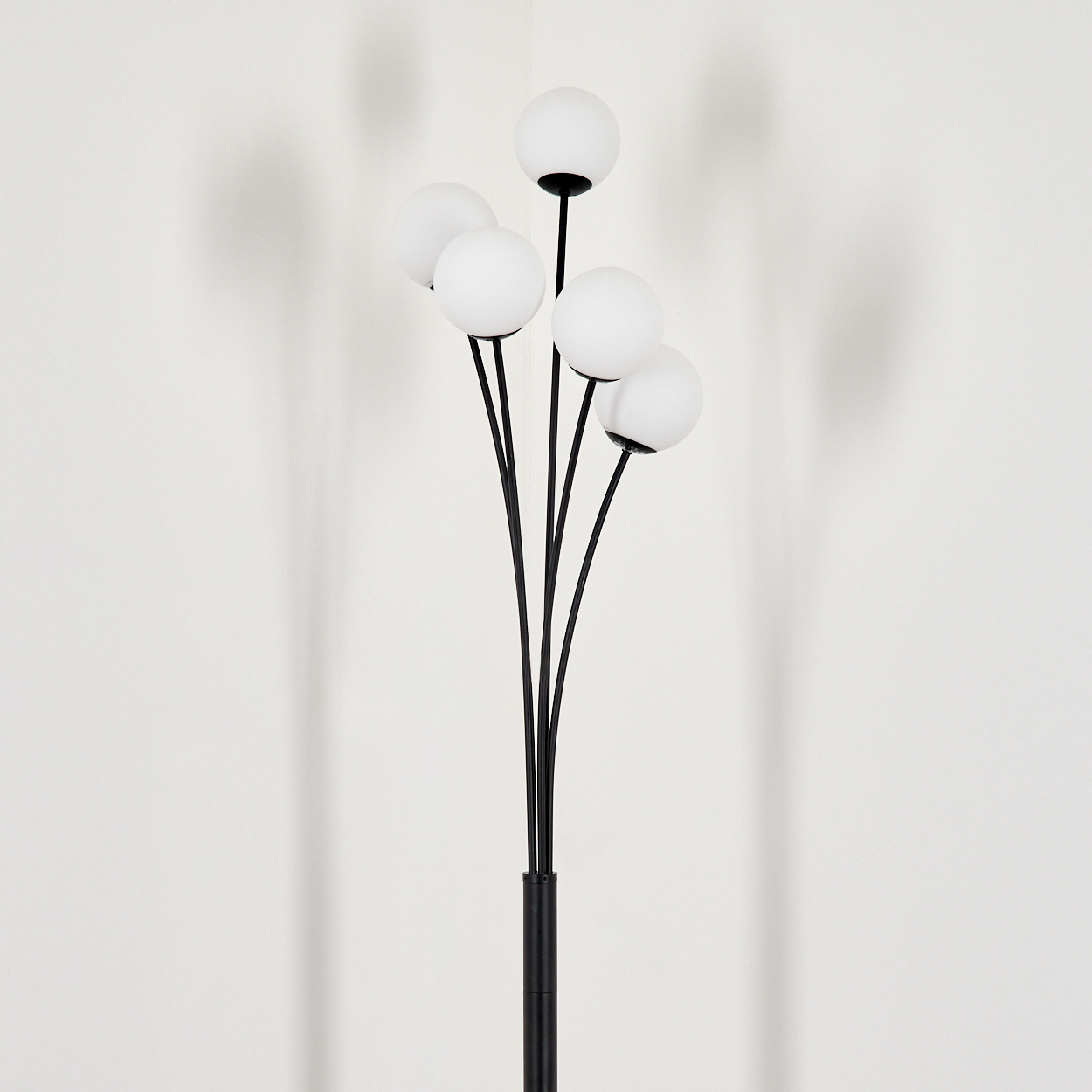 Eine schwarze Stehlampe mit sechs weißen Kugelleuchten. Die Lampe ist vor einer weißen Wand zentriert und wirft einen Schatten.