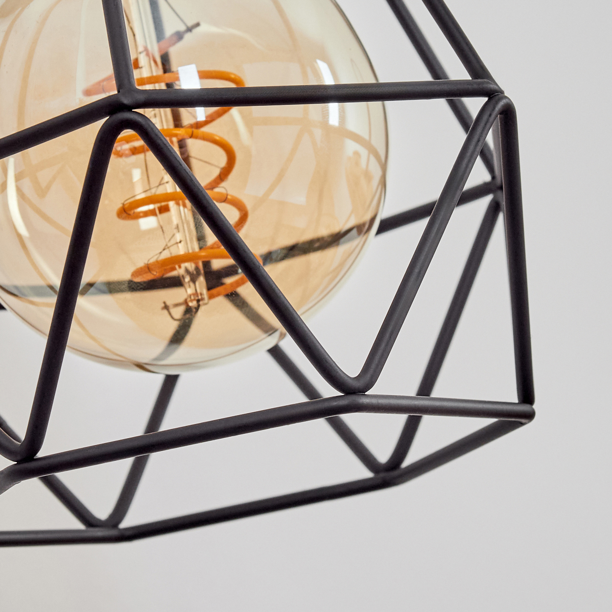 Nahaufnahme einer modernen Leuchte. Ein schwarzer, geometrischer Käfig umgibt eine bernsteinfarbene Glühbirne mit Spiralfaden vor weißem Hintergrund.
