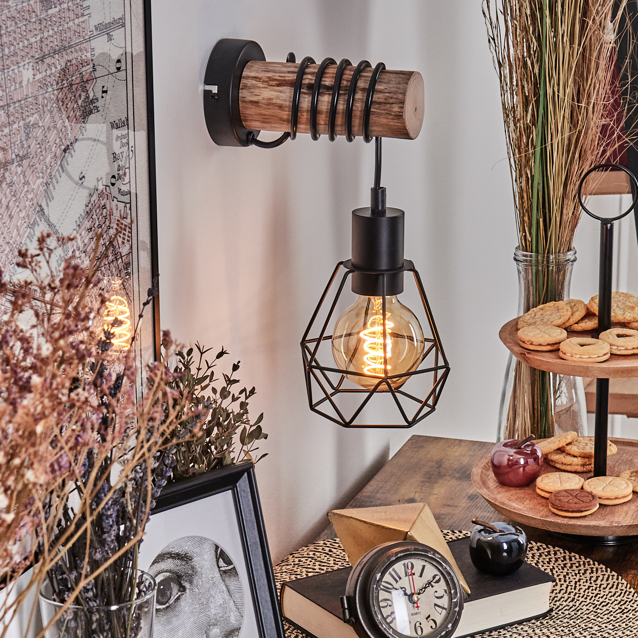 Eine Wandlampe mit schwarzem Metallkäfig und leuchtender Glühbirne. Kekse, eine Uhr und ein Bilderrahmen sind im Rahmen. Getrocknete Pflanzen.