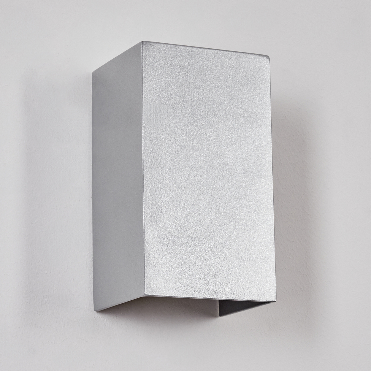 Eine silberne, rechteckige Wandleuchte ist an einer weißen Wand befestigt. Die Lampe ist metallisch und kastenförmig, mit offenem Boden.