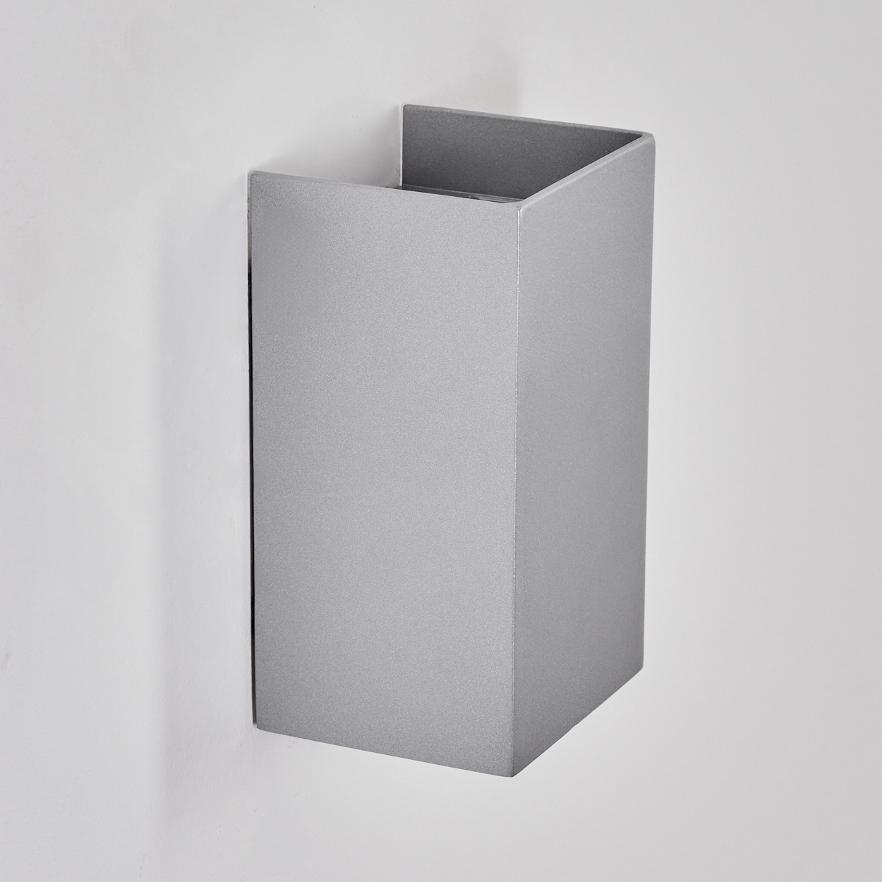 Eine silberne, rechteckige Wandleuchte an einer weißen Wand. Die Leuchte ist oben offen und hat ein minimalistisches Design.