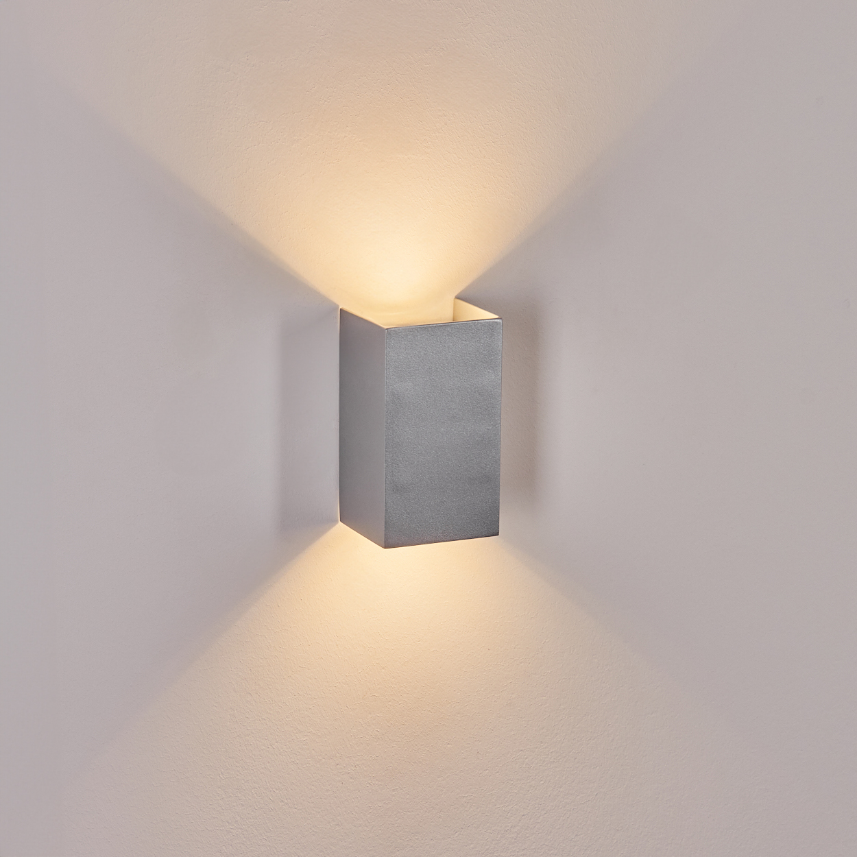 Eine silberne, würfelförmige Wandleuchte strahlt nach oben und unten Licht aus und wirft Schatten auf eine hellfarbige Wand.