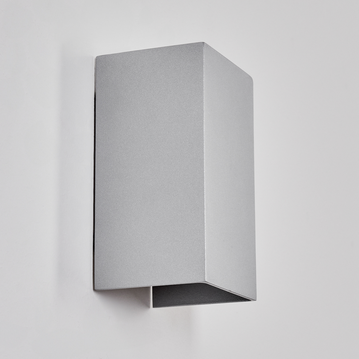 Eine silberne, rechteckige Wandleuchte ist an einer weißen Wand befestigt. Der untere Teil der Lampe ist dunkler.
