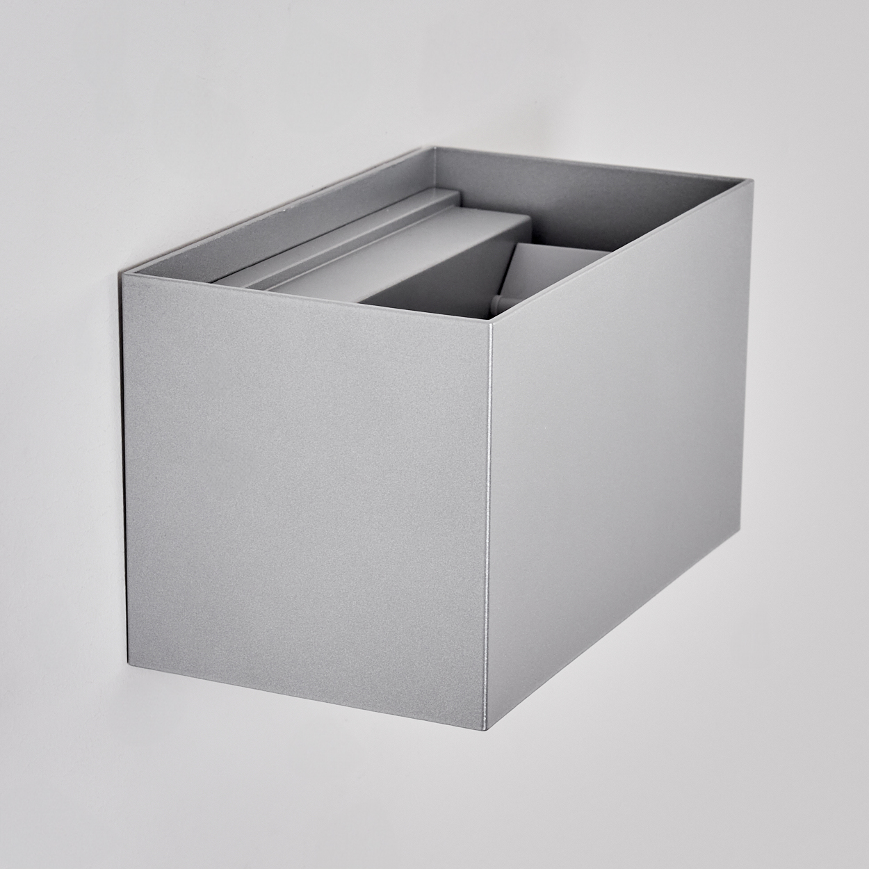 Eine silberne, rechteckige Leuchte ist an einer weißen Wand montiert. Die Leuchte hat ein geometrisches Design mit offener Oberseite.