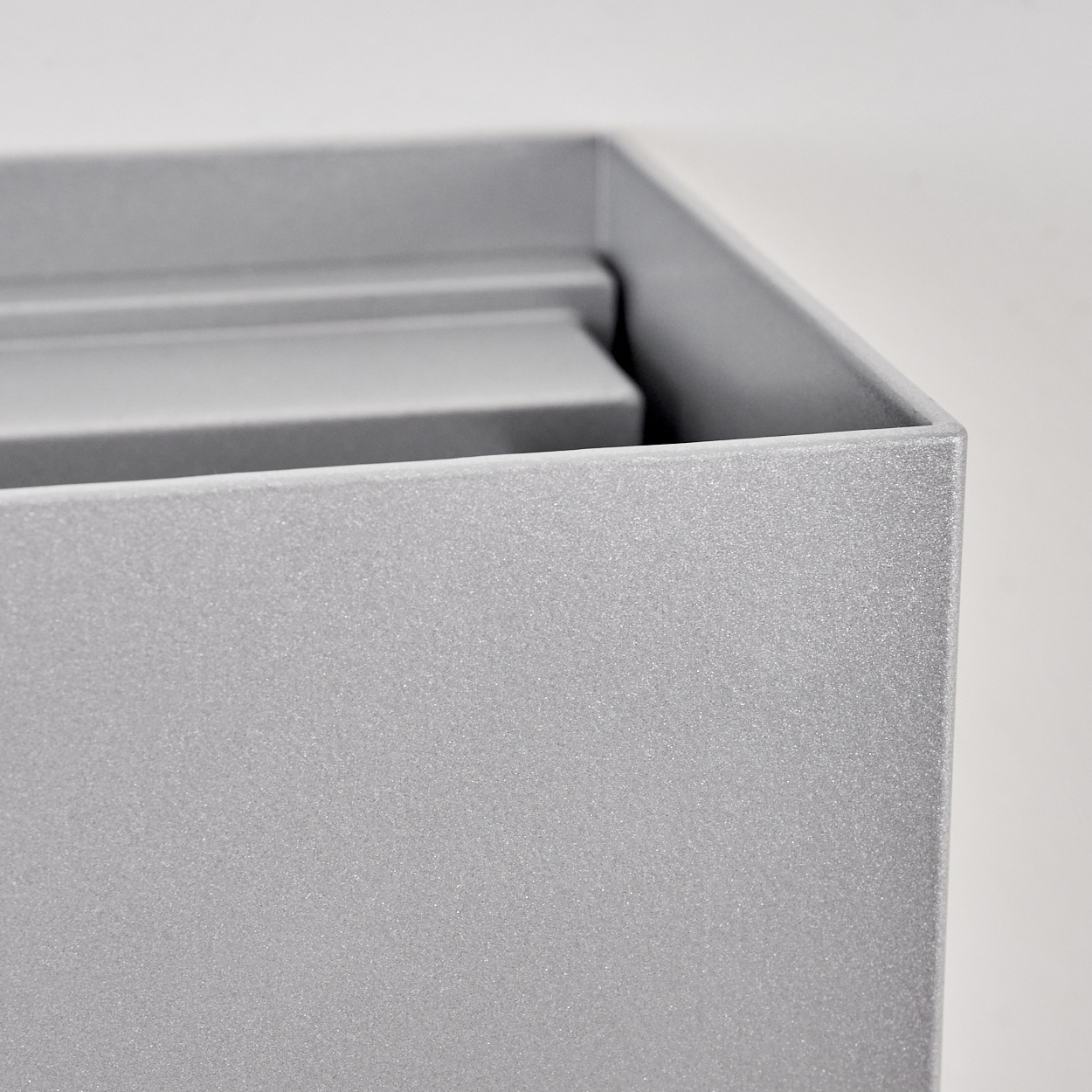 Nahaufnahme einer grauen rechteckigen Box mit strukturierter Oberfläche. Eine weitere Box ist teilweise sichtbar. Der Hintergrund ist weiß.