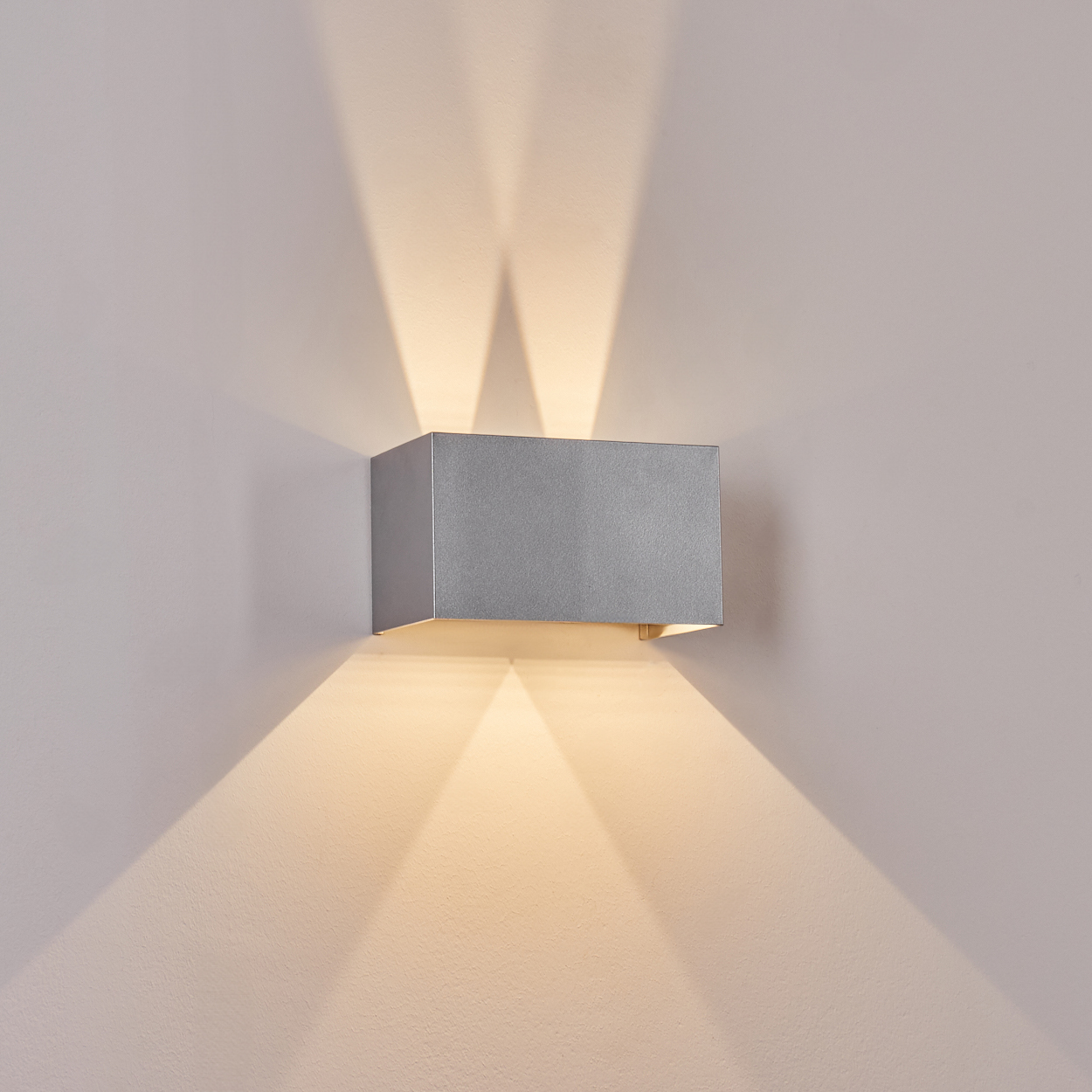 Eine silberne, rechteckige Wandleuchte ist an einer hellgrauen Wand angebracht. Licht strahlt nach oben und unten und wirft Schatten.