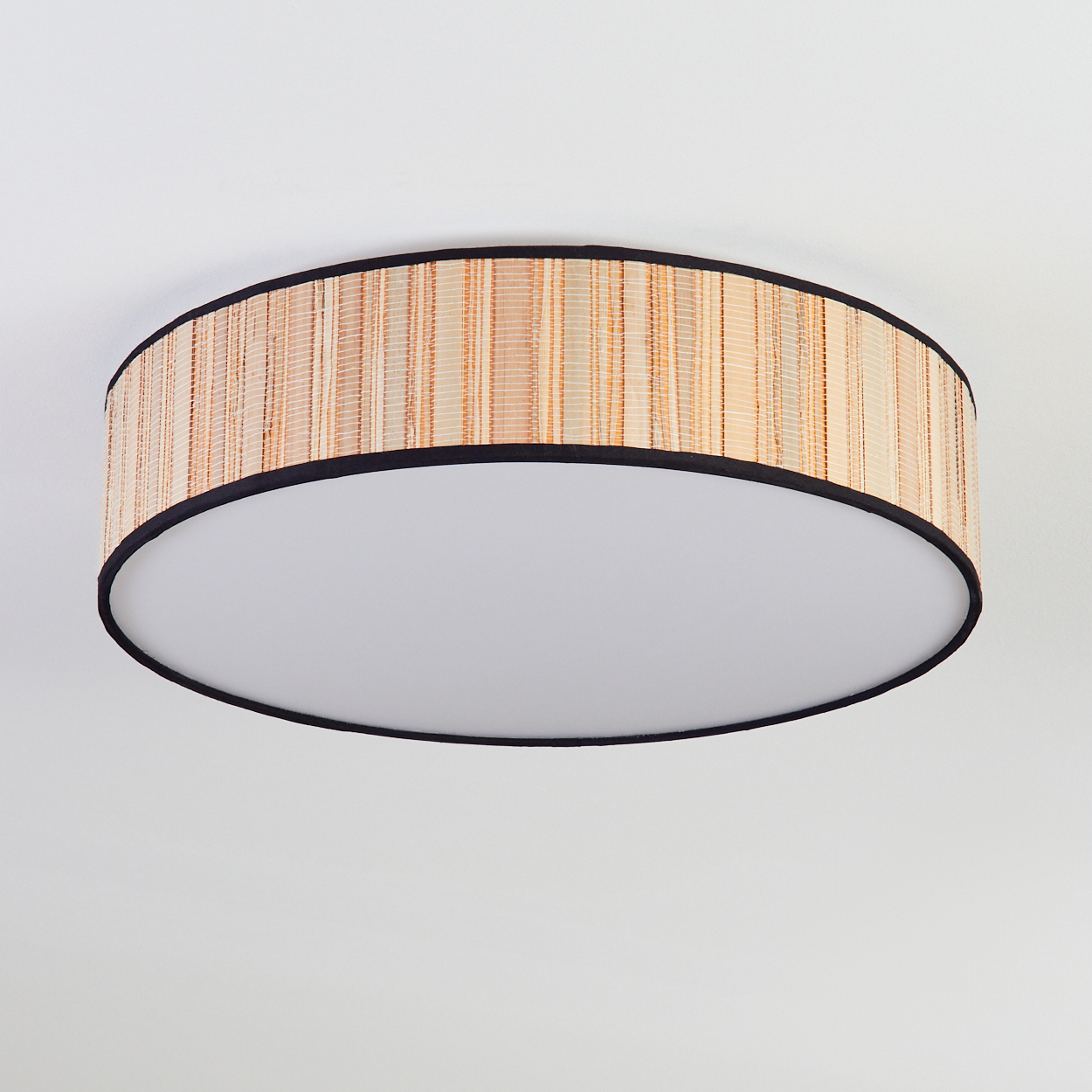 Eine runde Deckenlampe mit gewebtem Bambusschirm und schwarzem Rand, weißem Diffusor, vor weißer Decke.