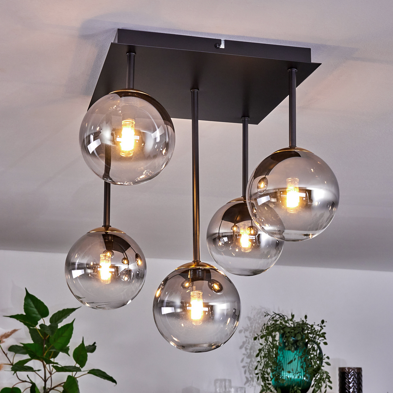 Moderner Kronleuchter mit fünf kugelförmigen Lampen, schwarzem Rahmen und goldenen Details. Pflanzen im Hintergrund, vor weißer Decke.