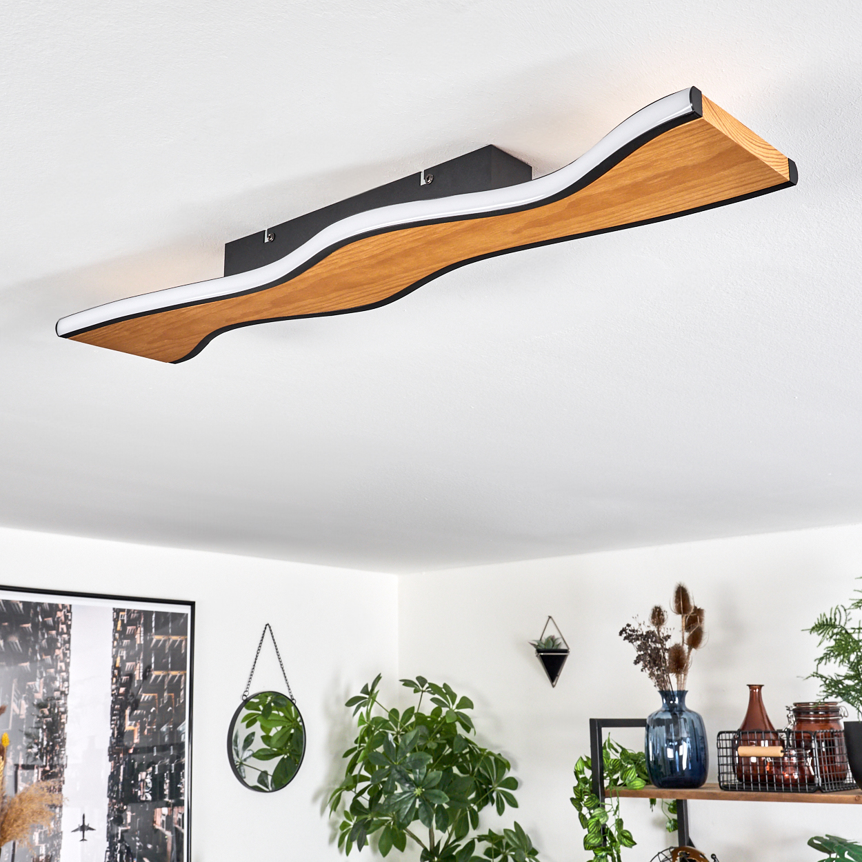 Eine moderne Deckenlampe aus Holz mit wellenförmigem Design ist an einer weißen Decke montiert. Pflanzen und Dekorationen sind im Hintergrund.