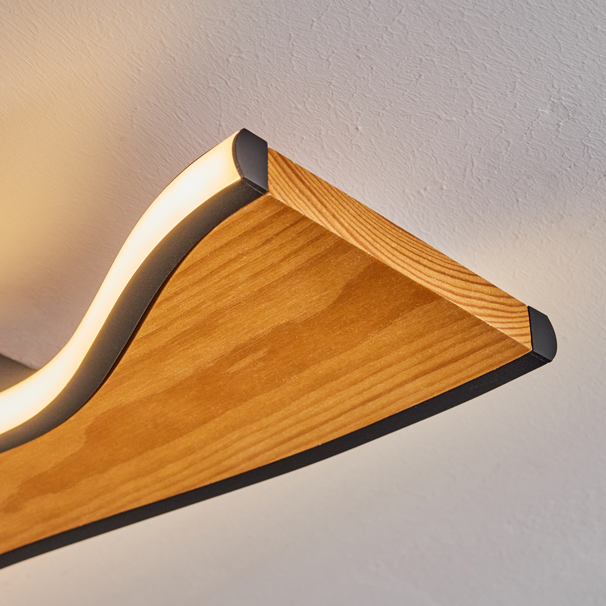 Holzleuchte mit schwarzem Rand. Das Licht kommt von unter der Holzkurve. Der Hintergrund ist eine strukturierte weiße Decke.