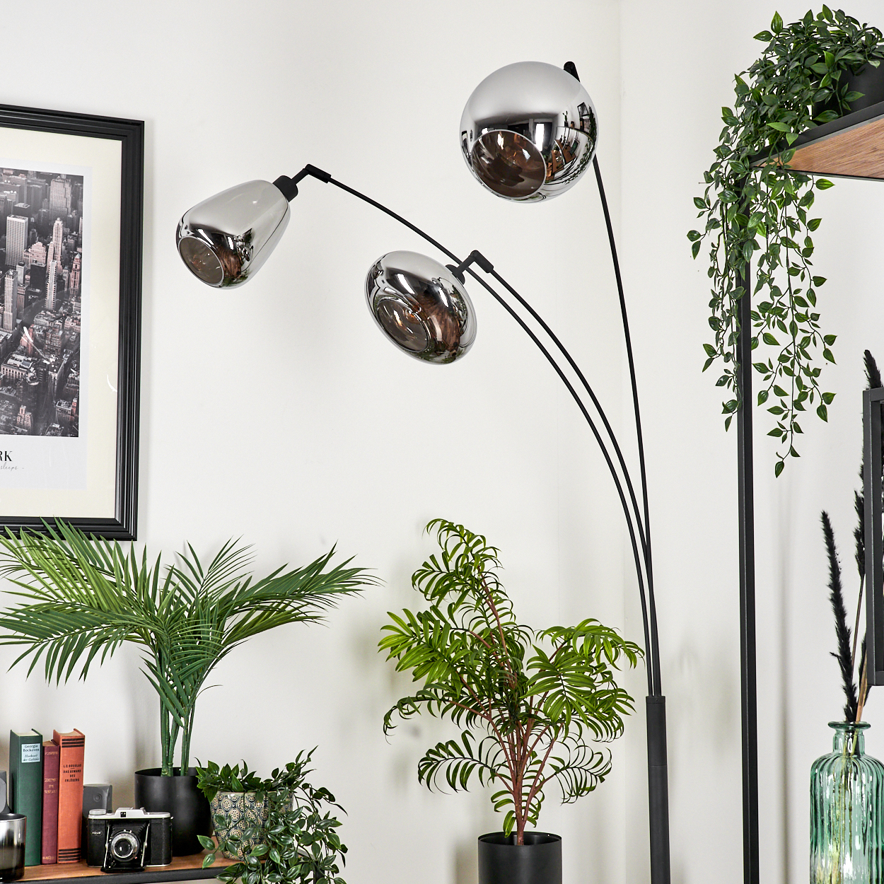 Eine moderne, schwarze Stehlampe mit drei Chromköpfen, umgeben von Topfpflanzen und einem schwarz gerahmten Stadtbild an der Wand.