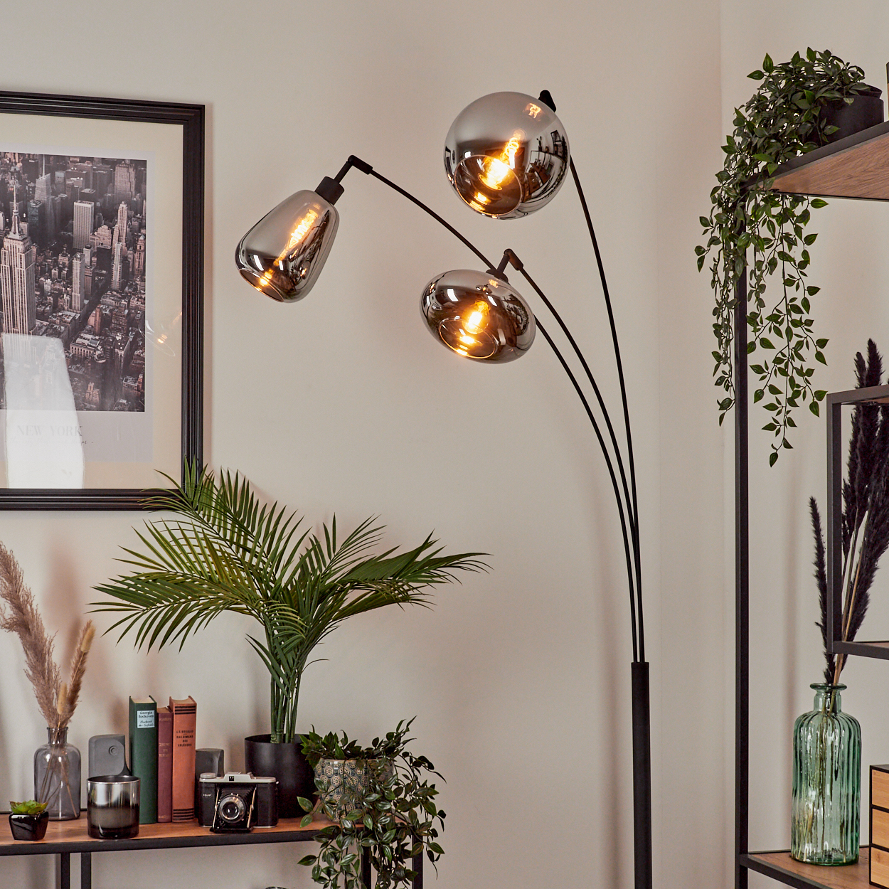 Eine moderne Stehlampe mit drei Glasschirmen. Der Raum hat Pflanzen, ein Regal, ein gerahmtes Stadtbild und einige Bücher.