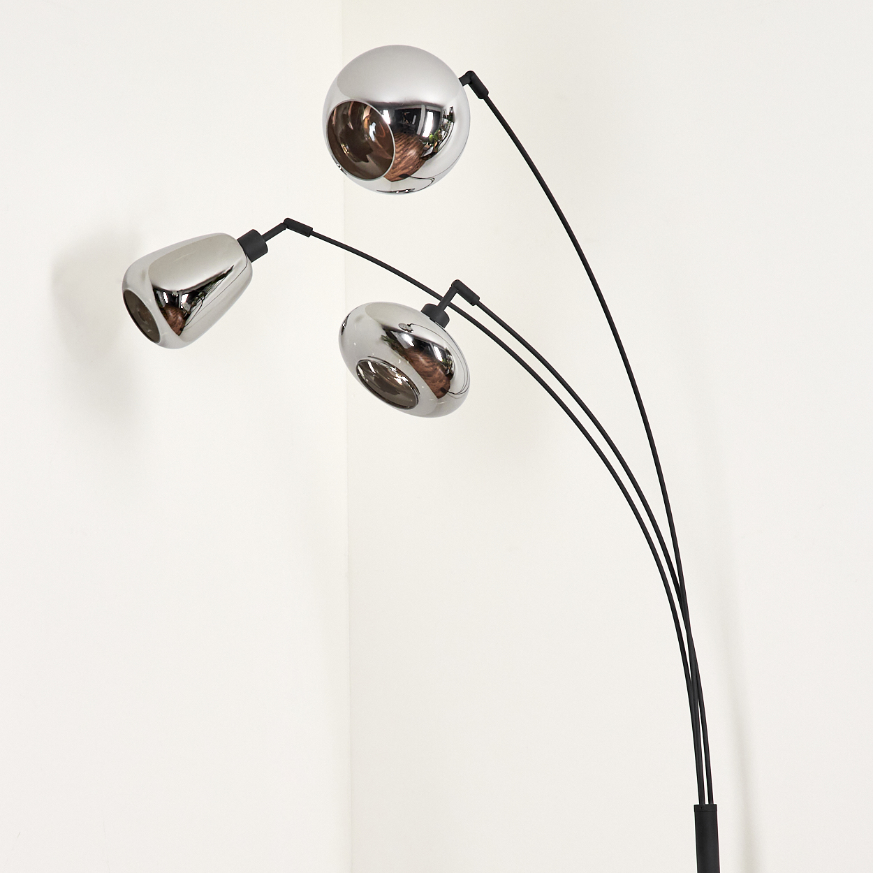 Eine moderne Stehlampe mit drei verchromten Kugellampen. Der Sockel ist schwarz und der Hintergrund ist eine weiße Ecke.