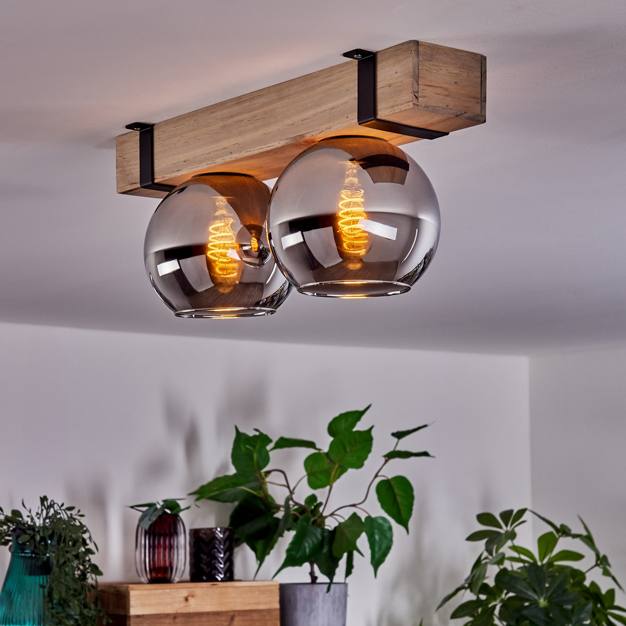 Eine Deckenleuchte mit zwei Glaskugeln, Holzbalken und schwarzen Metallhaltern. Pflanzen sind im Vordergrund. Das Licht ist an.