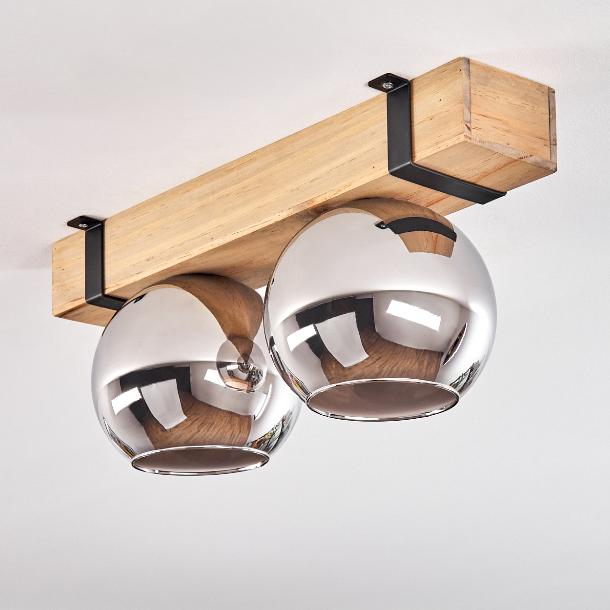 Eine Deckenleuchte mit zwei metallischen, kugelförmigen Lampen, die von einem Holzbalken abgehängt sind, der von schwarzen Metallbügeln getragen wird.