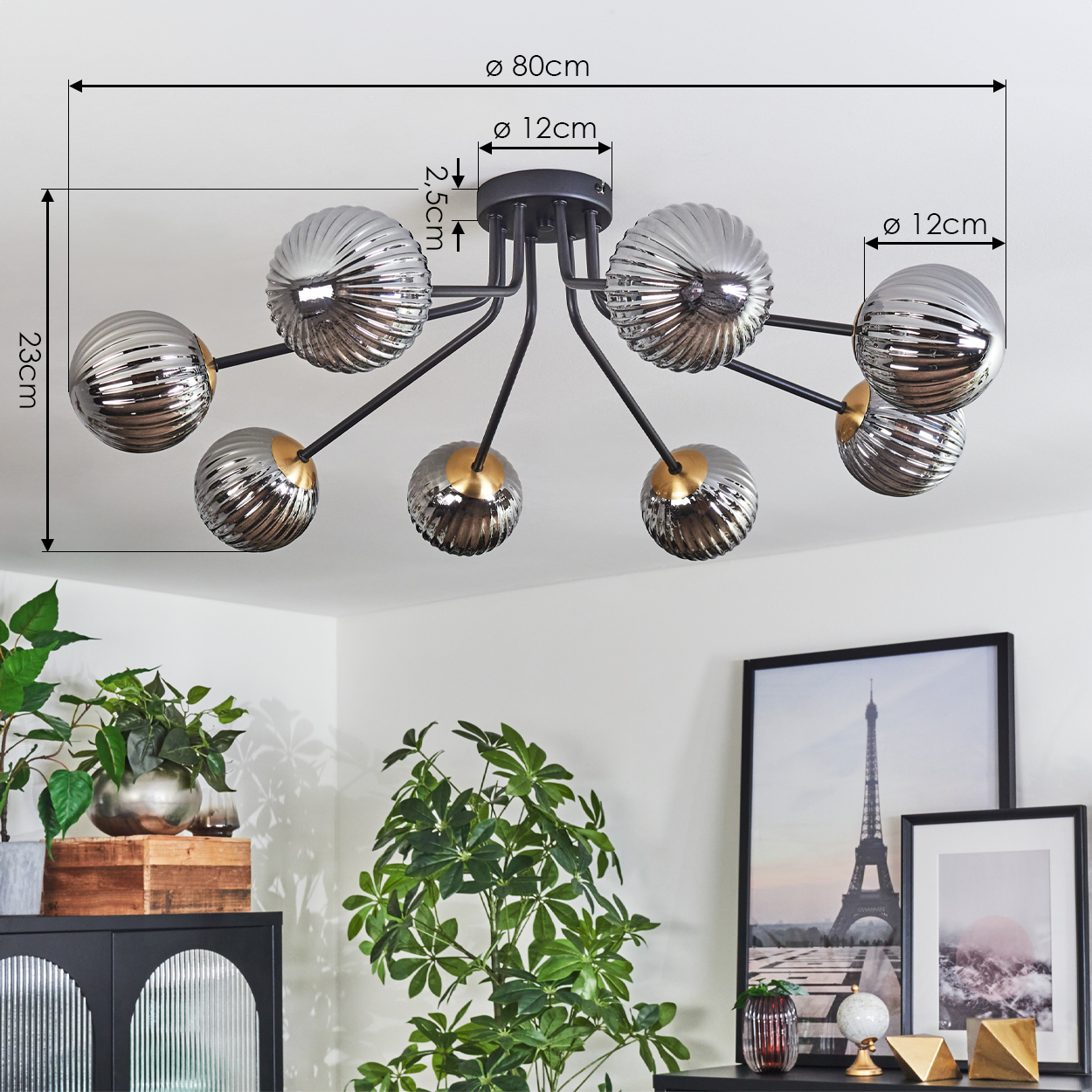 Eine moderne Deckenlampe mit acht Glaskugeln. Die Lampe ist schwarz mit goldenen Akzenten. Pflanzen und gerahmte Bilder darunter.