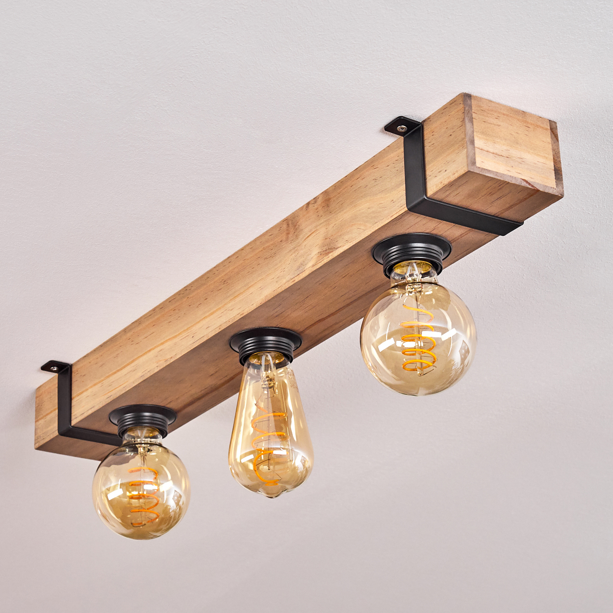 Ein Holzbalken mit drei Glühbirnen. Die Glühbirnen sind bernsteinfarben, und der Balken ist mit schwarzen Metallhalterungen an der Decke befestigt.