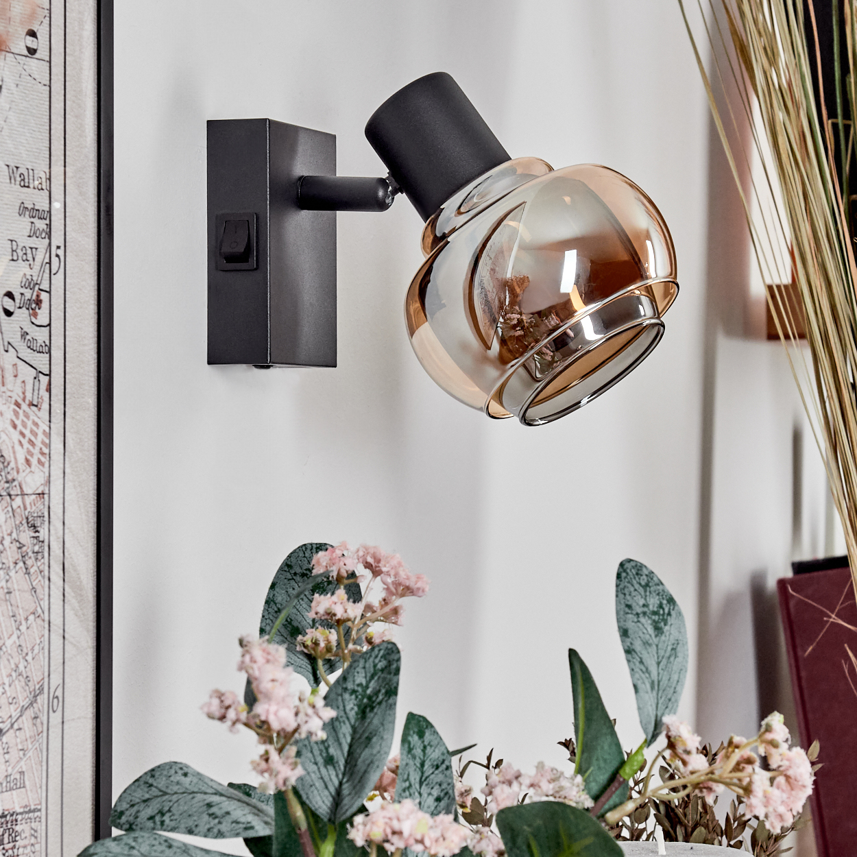 Eine schwarze Wandleuchte mit Glasschirm neben einer Karte und Blumen. Das Licht ist nach unten gerichtet. Die Szene befindet sich an einer weißen Wand.