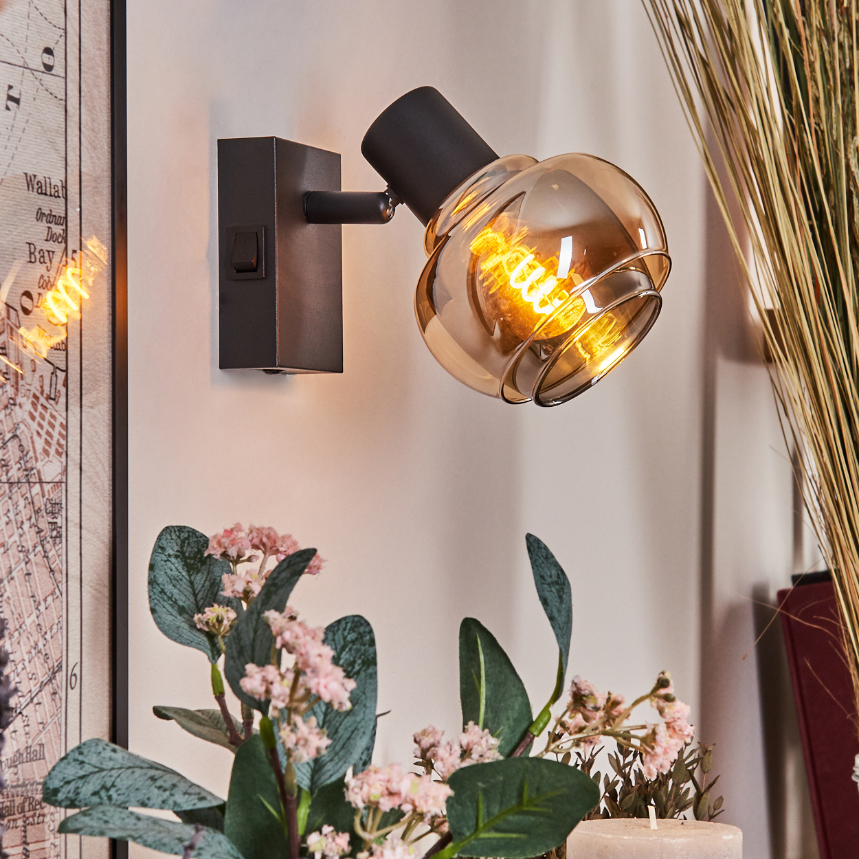 Eine schwarze Wandlampe mit Glasschirm ist an der Wand montiert. Blumen und eine Kerze sind im Vordergrund. Links ist eine Karte.