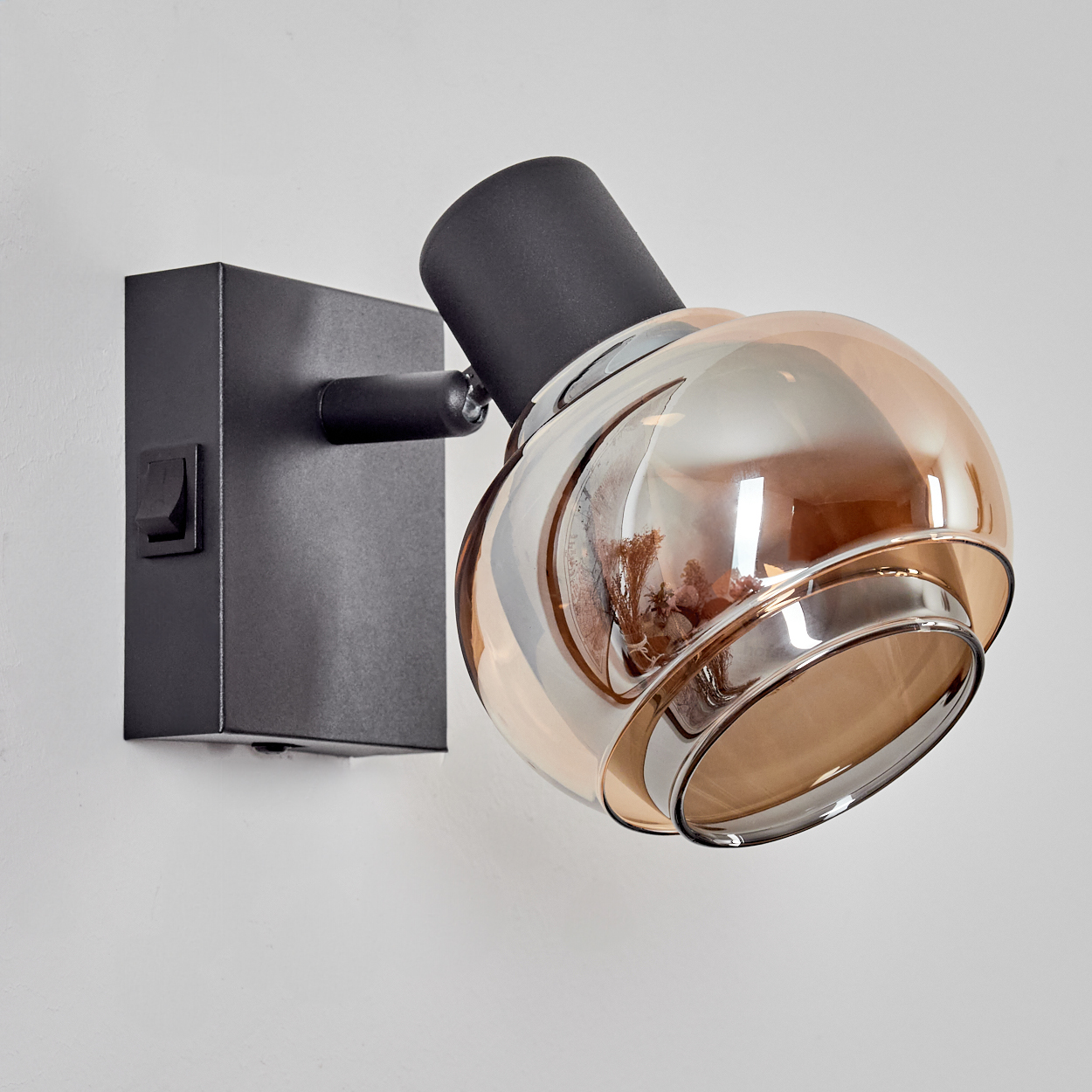 Eine schwarze Wandlampe mit einem kugelförmigen bernsteinfarbenen Glasschirm ist an einer weißen Wand montiert. Die Lampe hat einen Schalter.