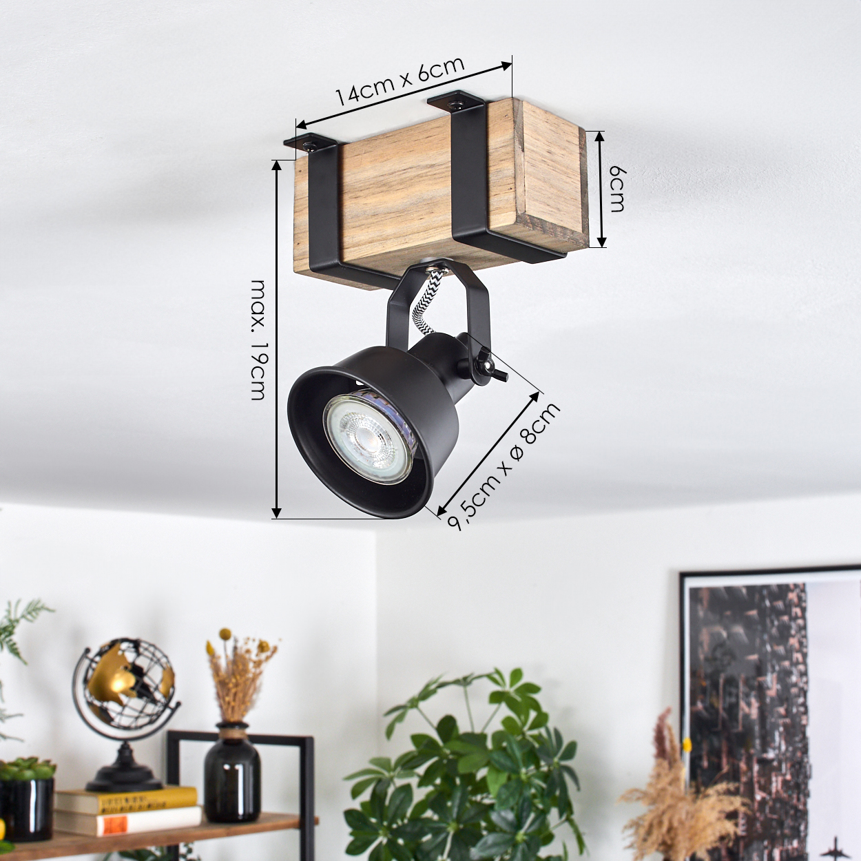 Schwarze Strahlerlampe auf Holzsockel an einer weißen Decke montiert. Der Raum hat Dekoration auf einem Regal und Pflanzen.
