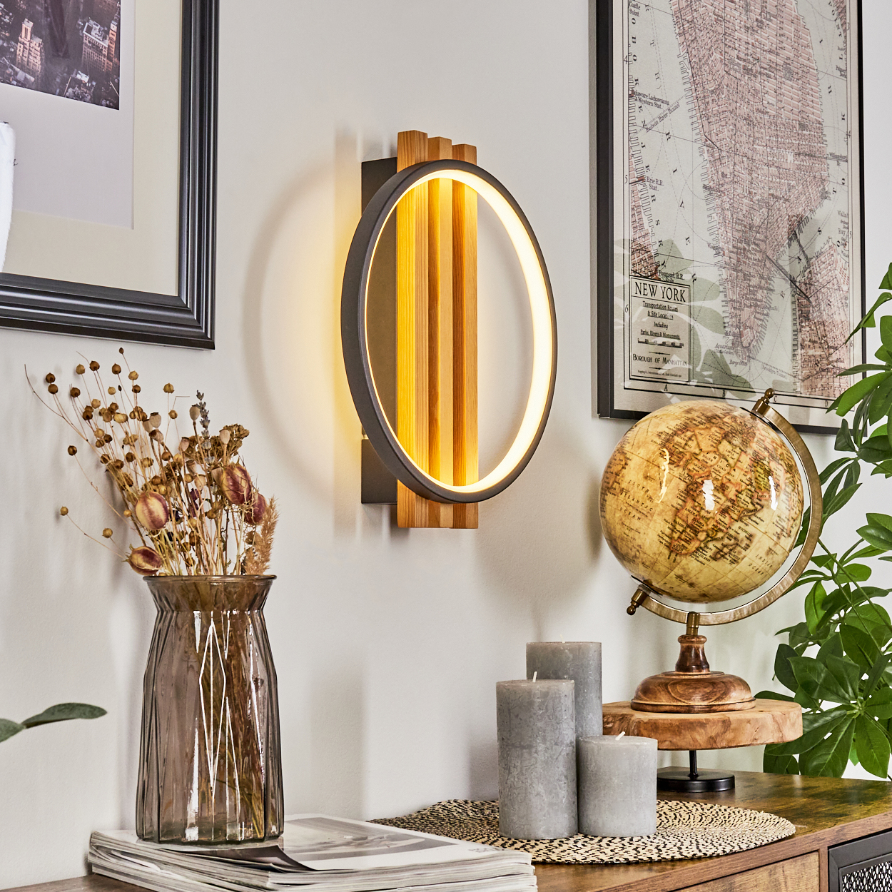 Eine moderne Wandlampe mit rundem Licht, ein Globus, Kerzen und eine Vase mit Trockenblumen stehen auf einem Holztisch.